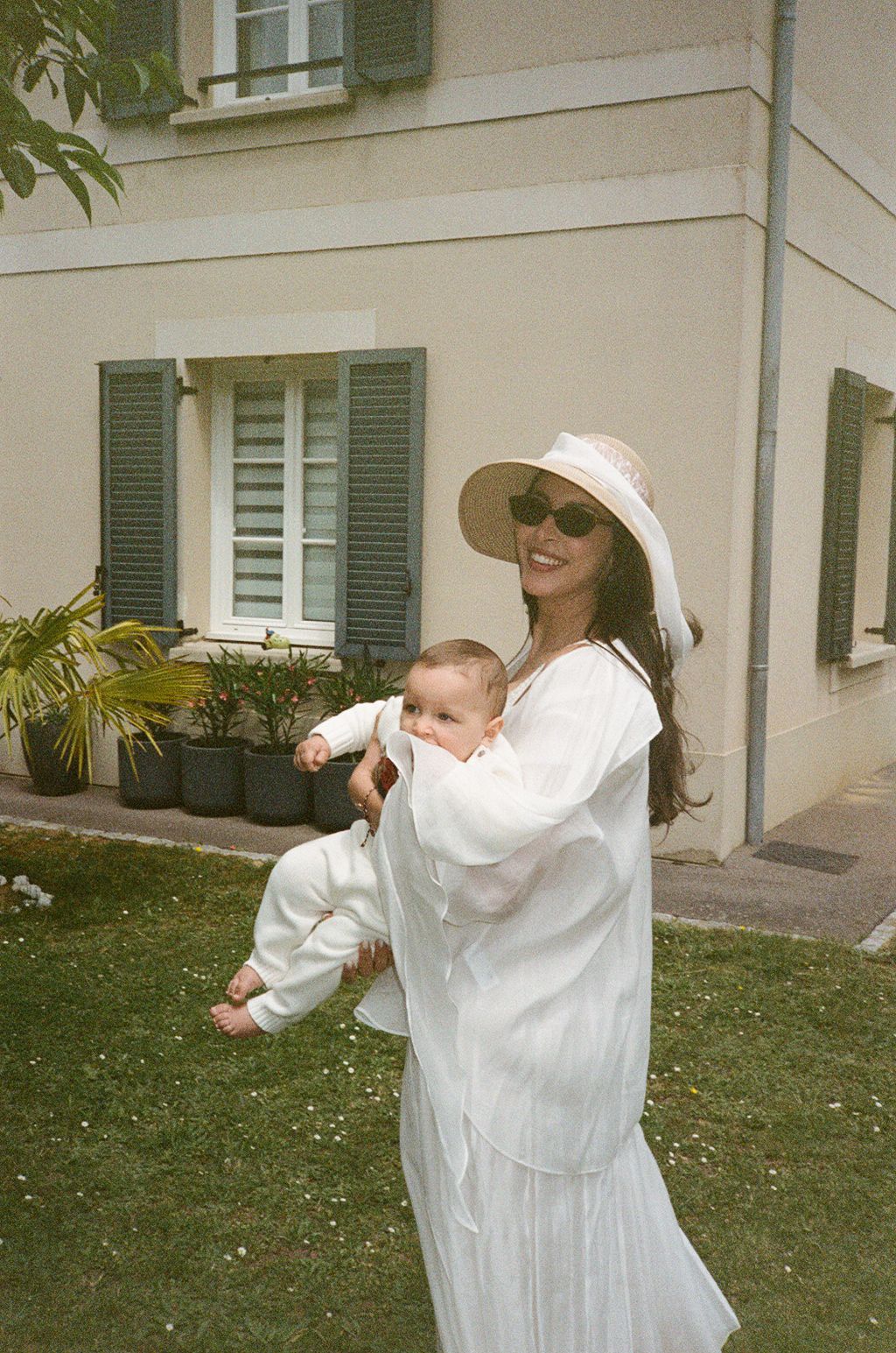 Woman in white dress and sun hat holding a baby outdoors in front of a French-style house with green shutters.
