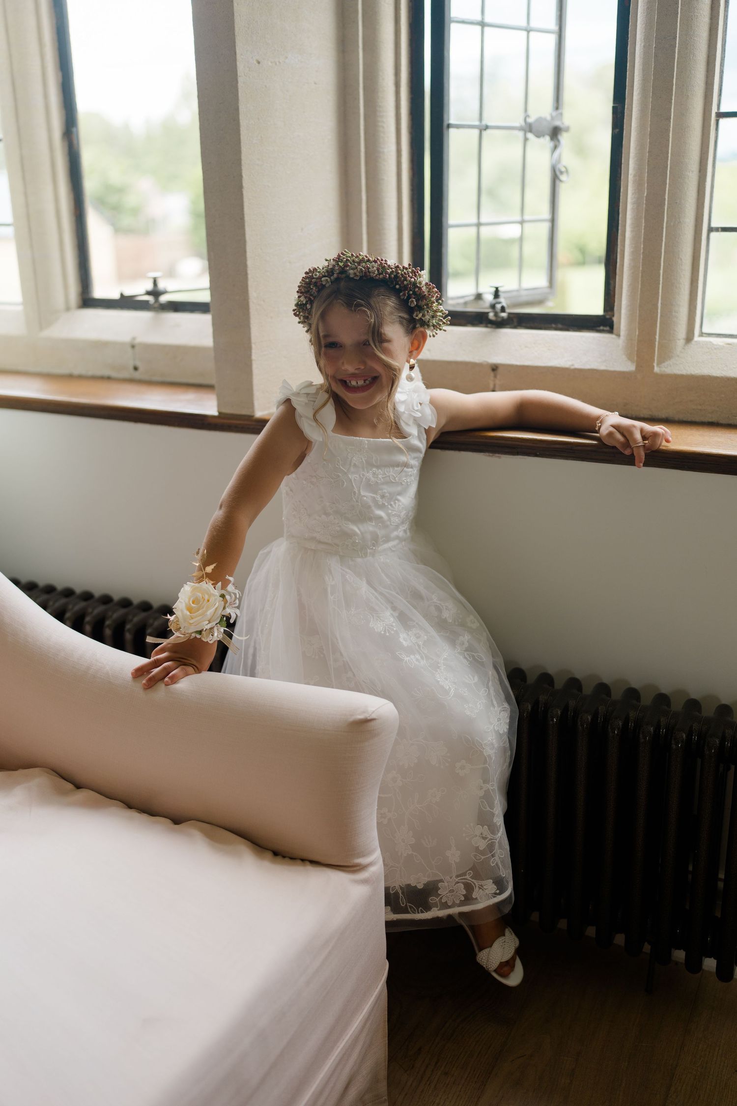 Joyful young flower girl in white dress with floral crown playing near window seat indoors.