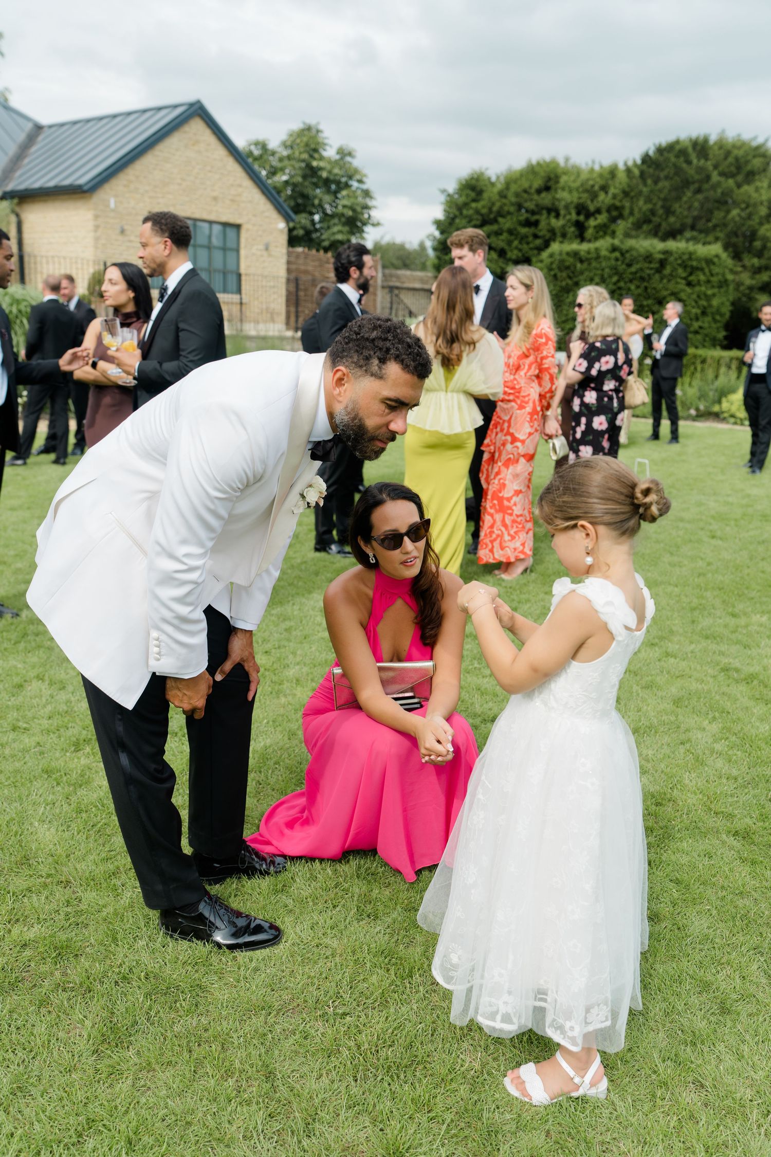 Groom in white tuxedo jacket bends to greet flower girl in white dress at outdoor wedding reception.