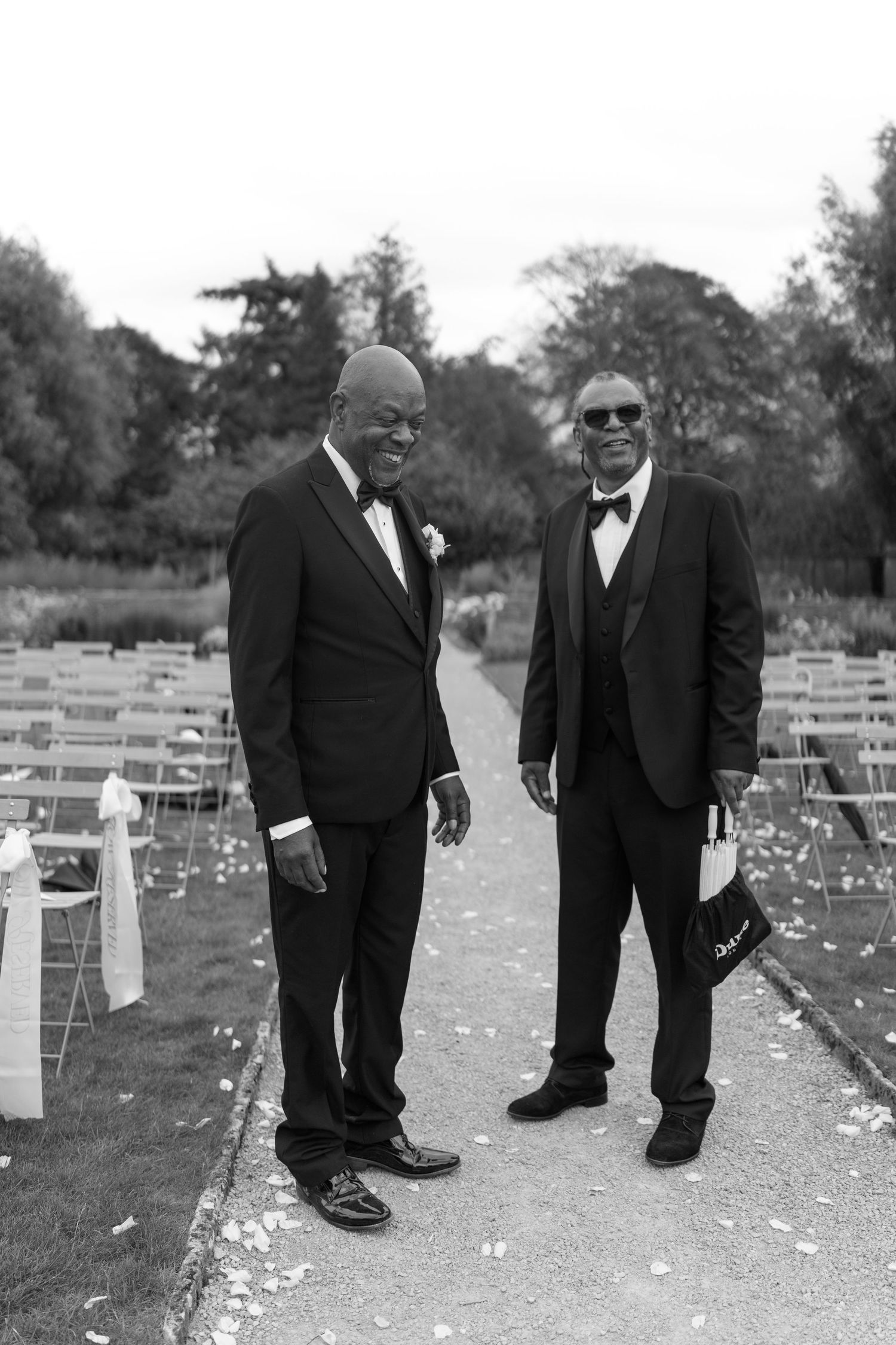 Two elegantly dressed men in black tuxedos smiling at an outdoor wedding ceremony in black and white.