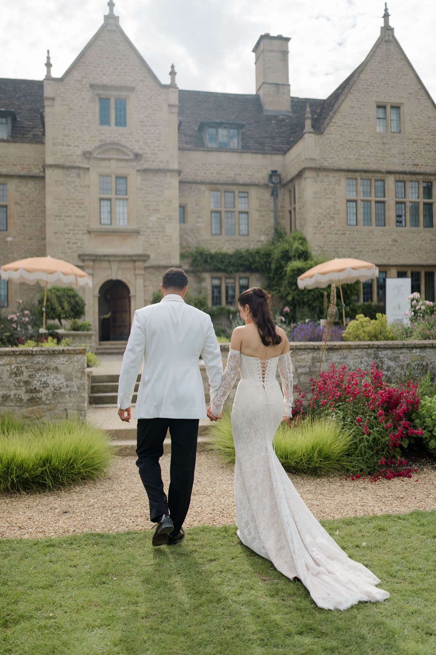 Bride in lace off-shoulder gown and groom in white jacket walk hand in hand toward stone manor house.