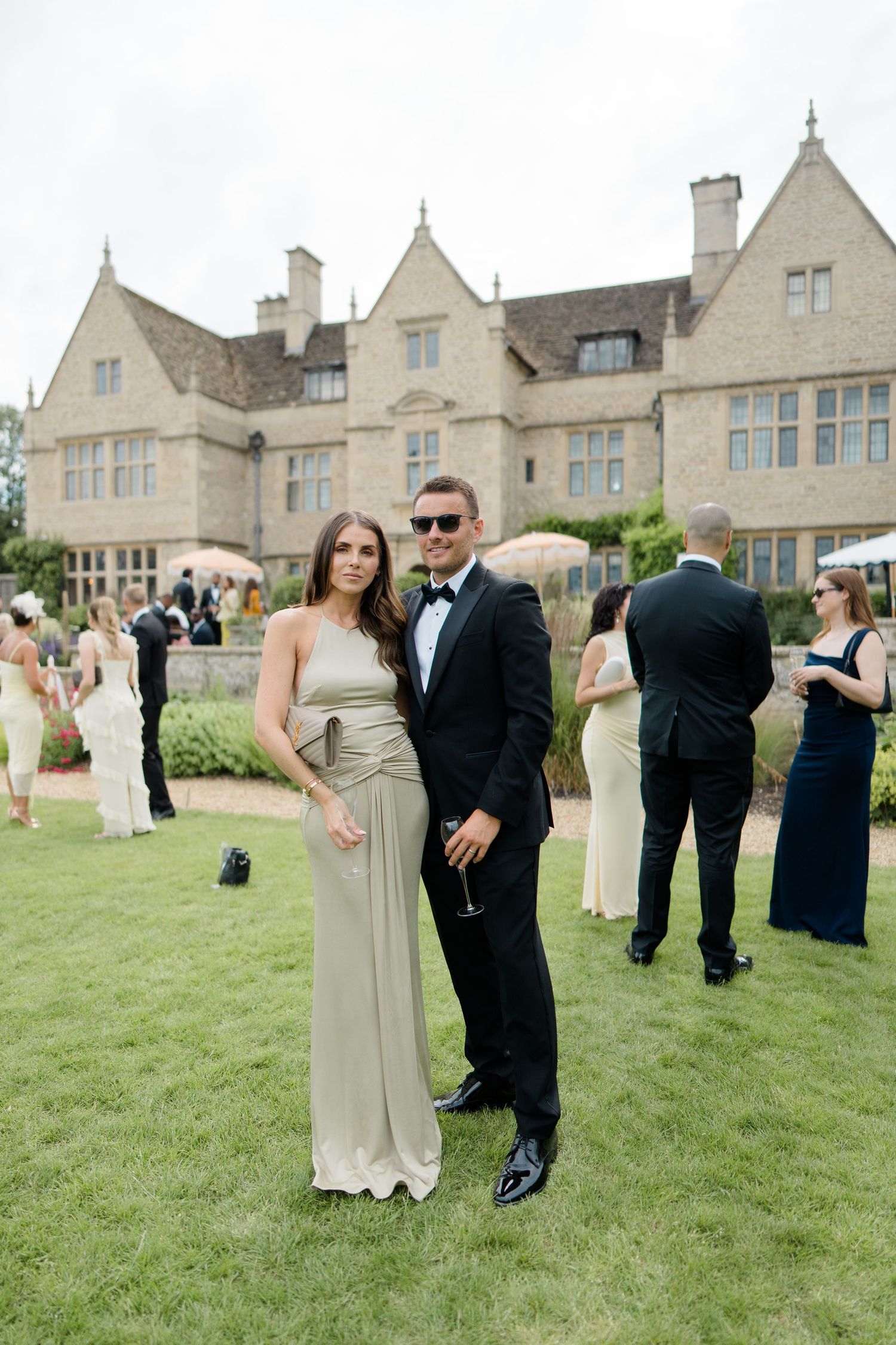 Elegant couple in formal attire posing on lawn outside a grand stone manor house at a wedding reception.