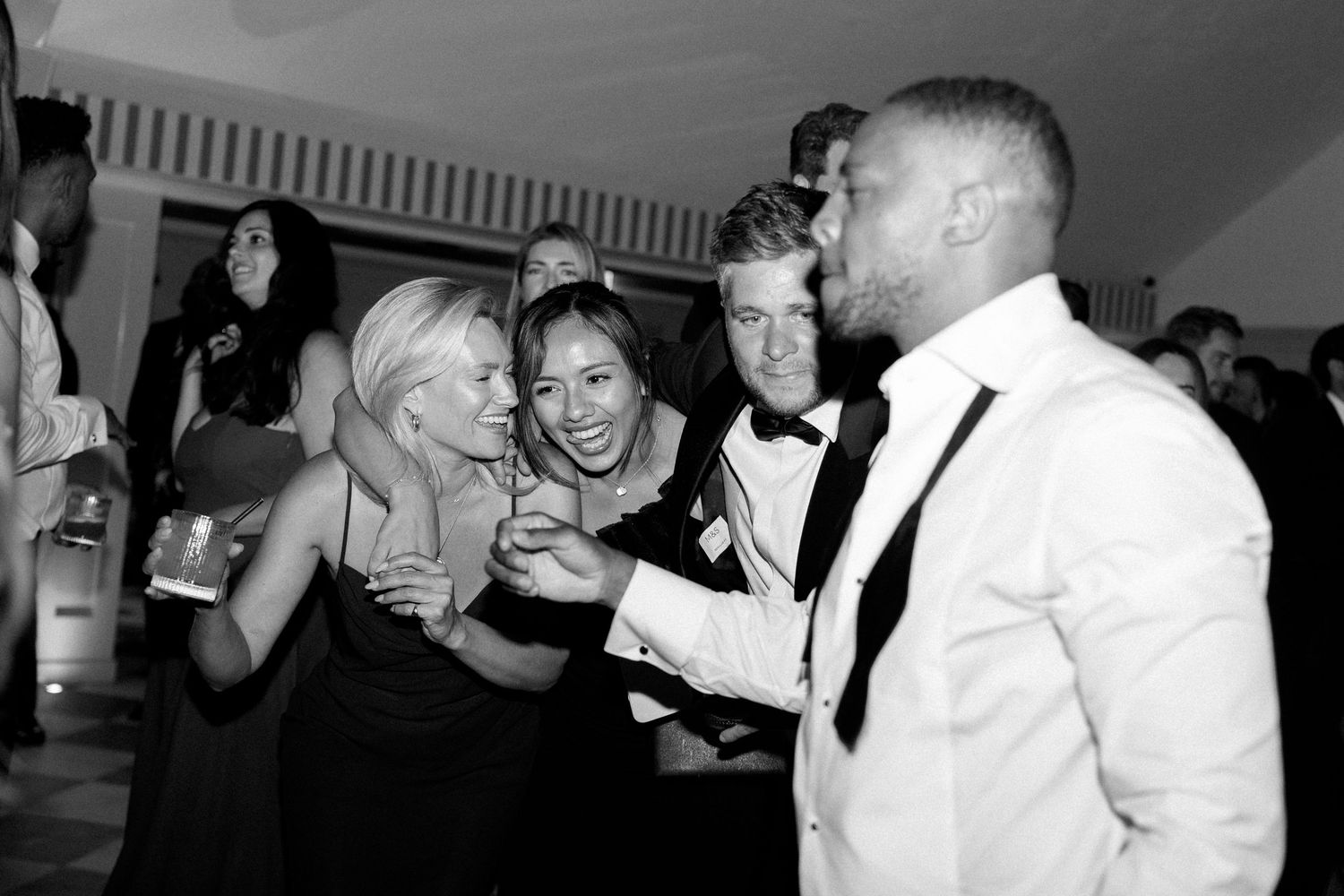 Black and white photo of guests laughing with groom in white tuxedo at wedding reception.