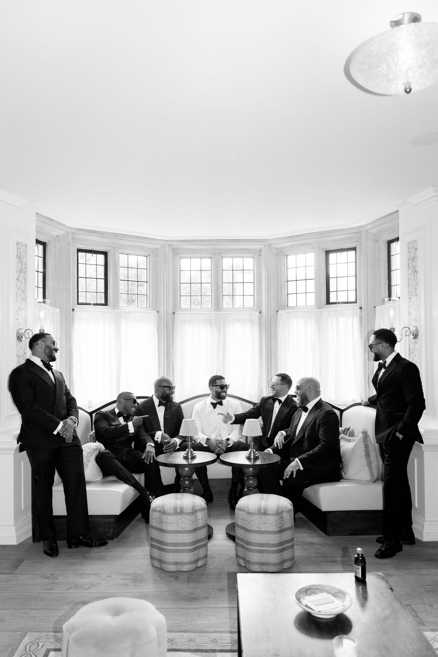 A black and white photo of groomsmen in tuxedos gathered in a bright bay window room before a wedding.