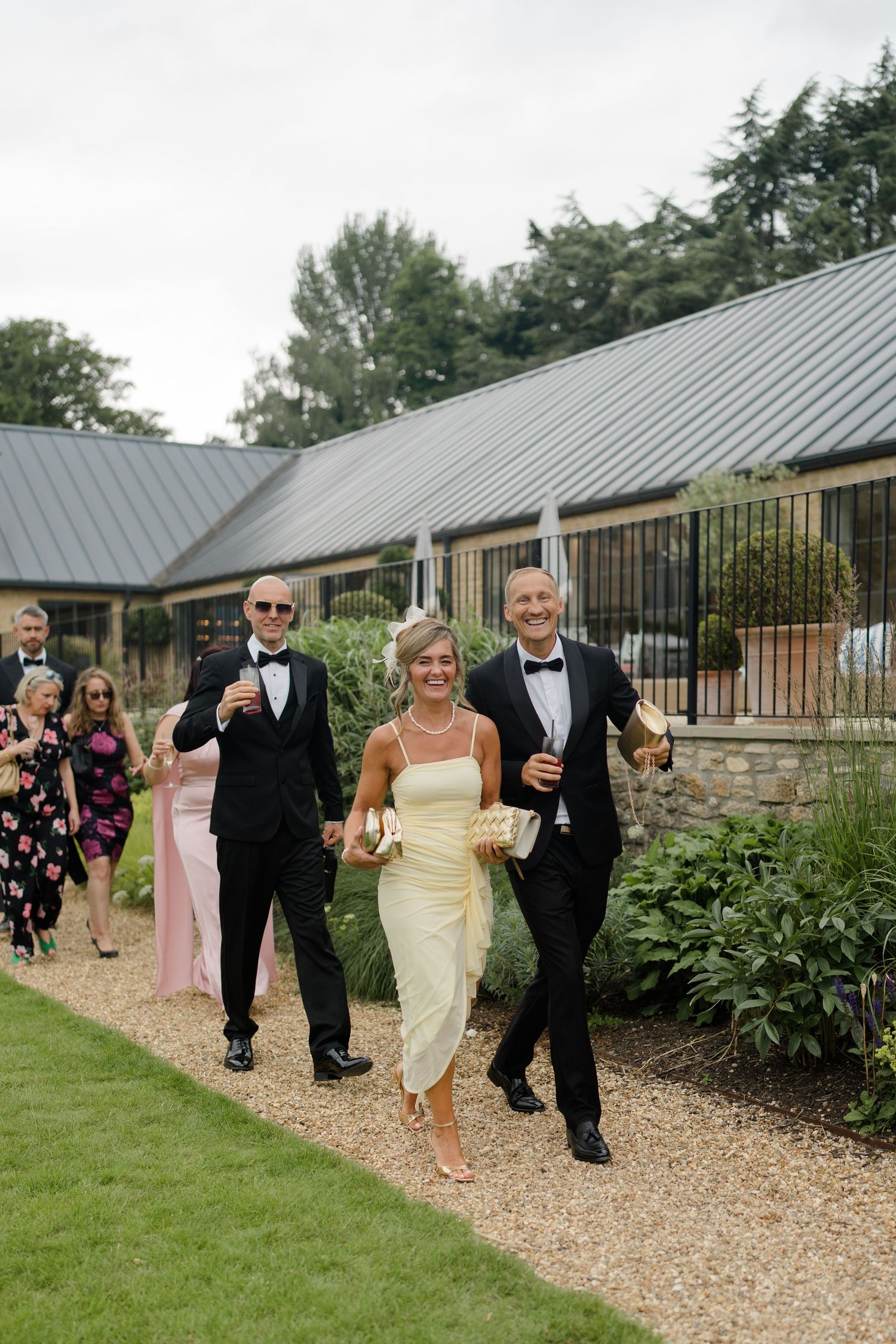 Wedding guests in formal attire walking along garden path at elegant venue.