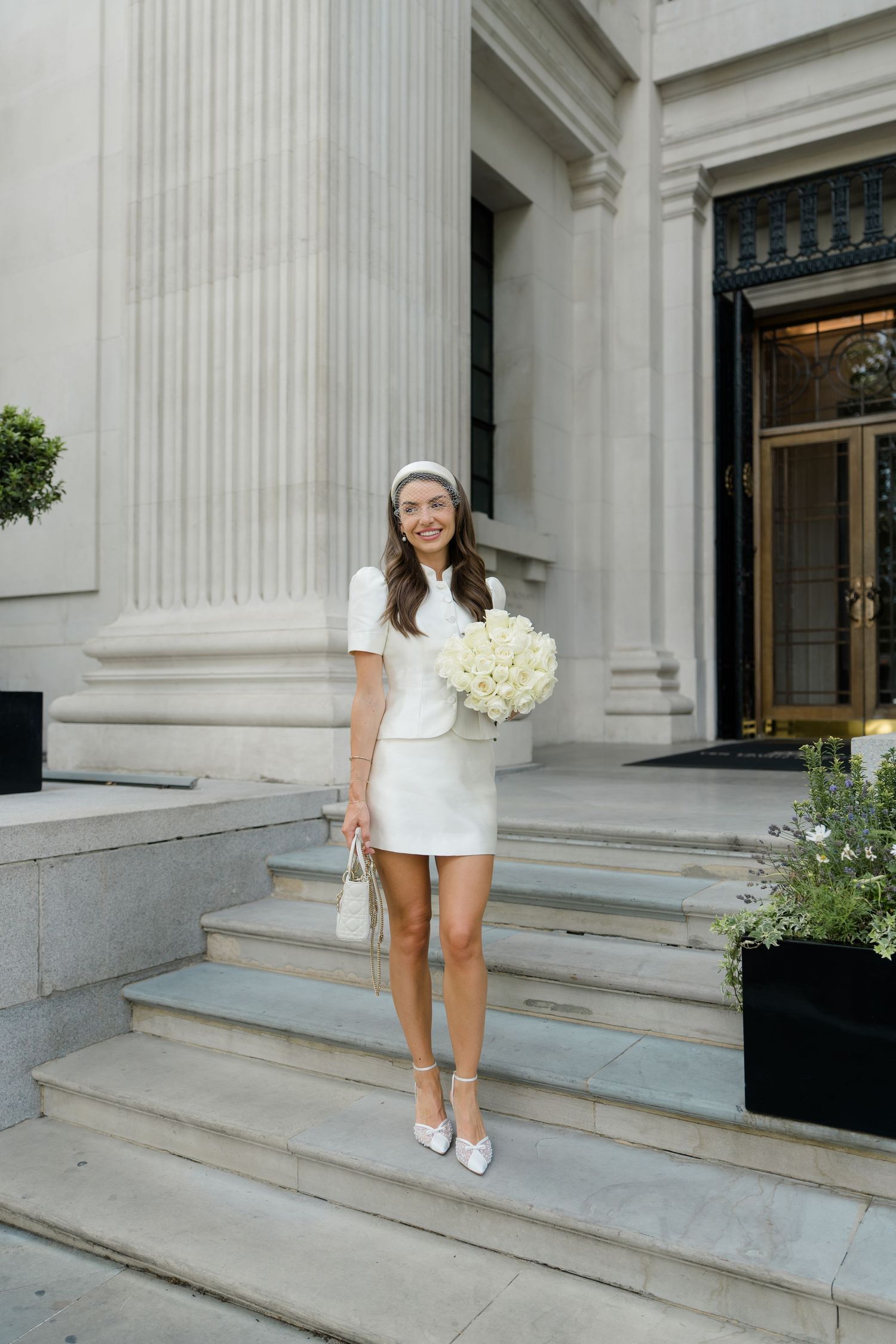 Bride in white mini dress and headband holding bouquet on stone steps of elegant building.