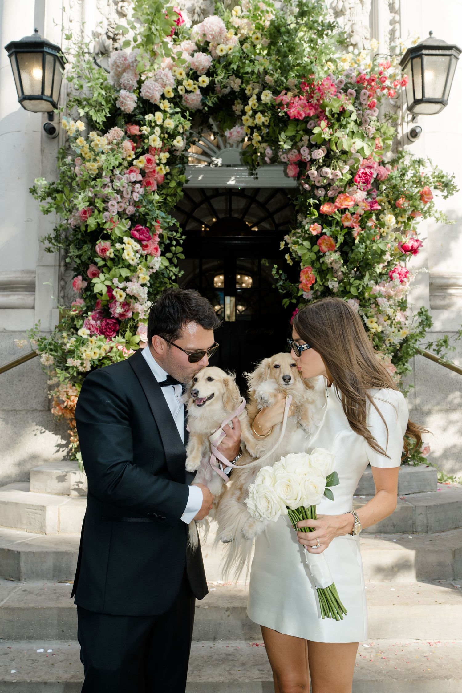 Bride and groom kissing their dogs in front of stunning floral wedding arch.