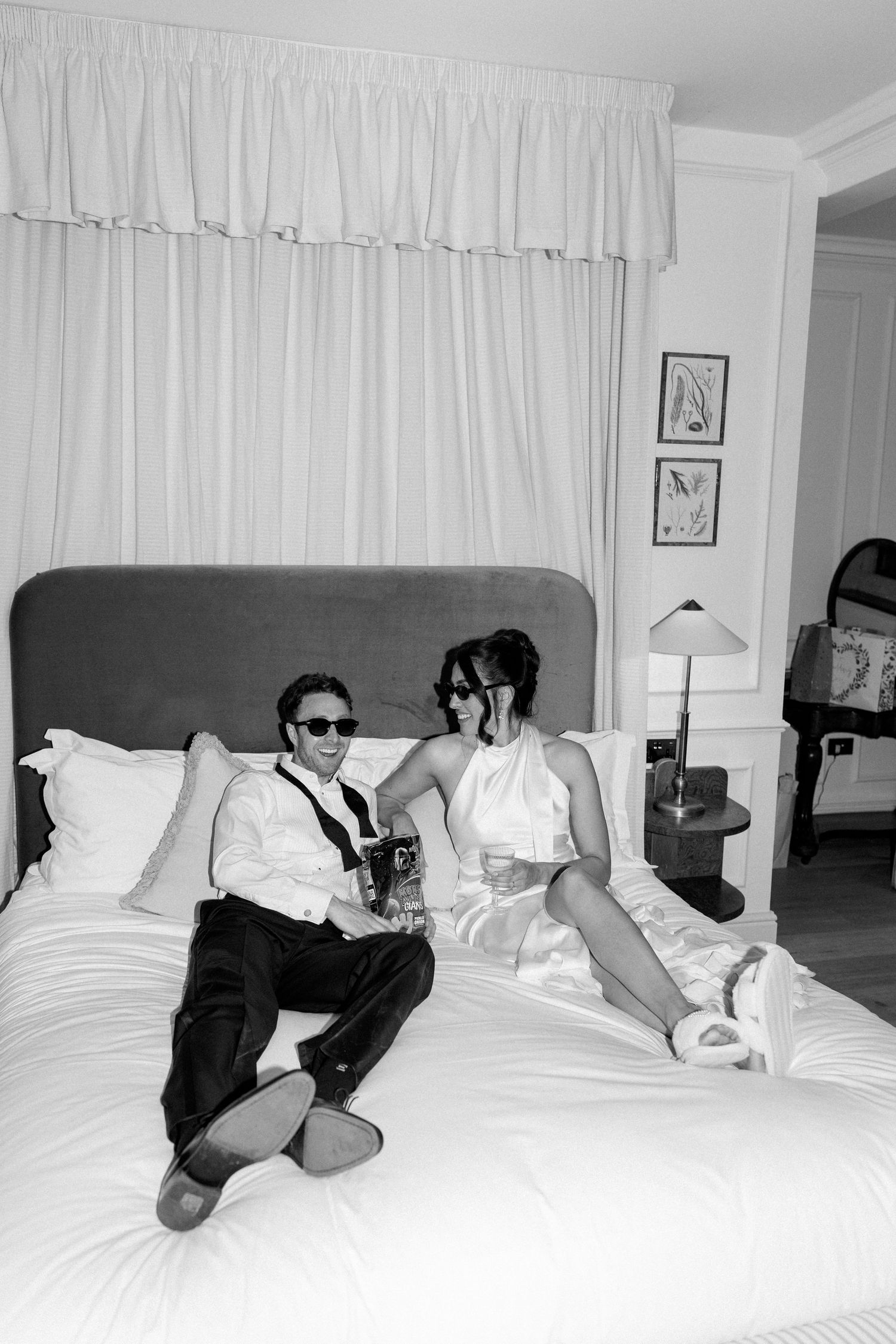 Black and white photo of bride and groom relaxing on hotel bed wearing sunglasses after wedding.