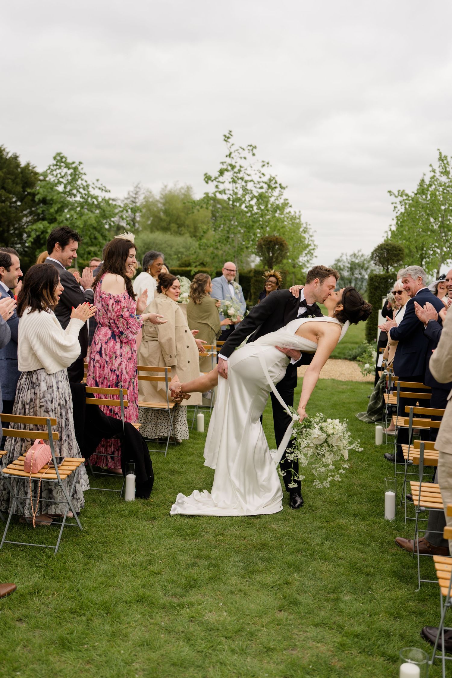 Groom dips bride for a kiss at outdoor wedding ceremony as guests applaud along the aisle.