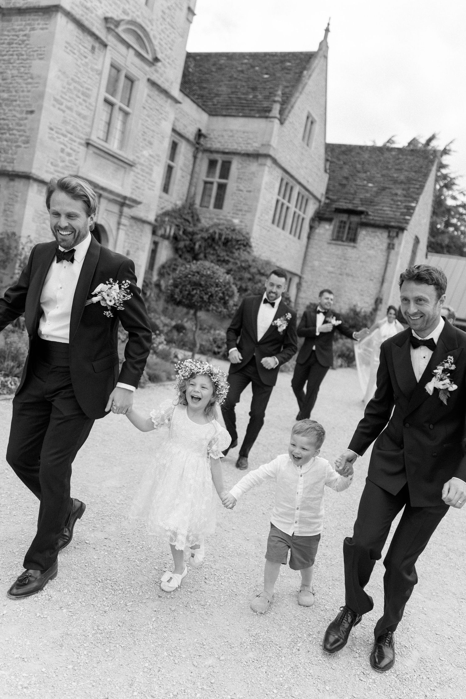 Black and white photo of groomsmen in tuxedos laughing with flower girl and page boy at manor wedding.
