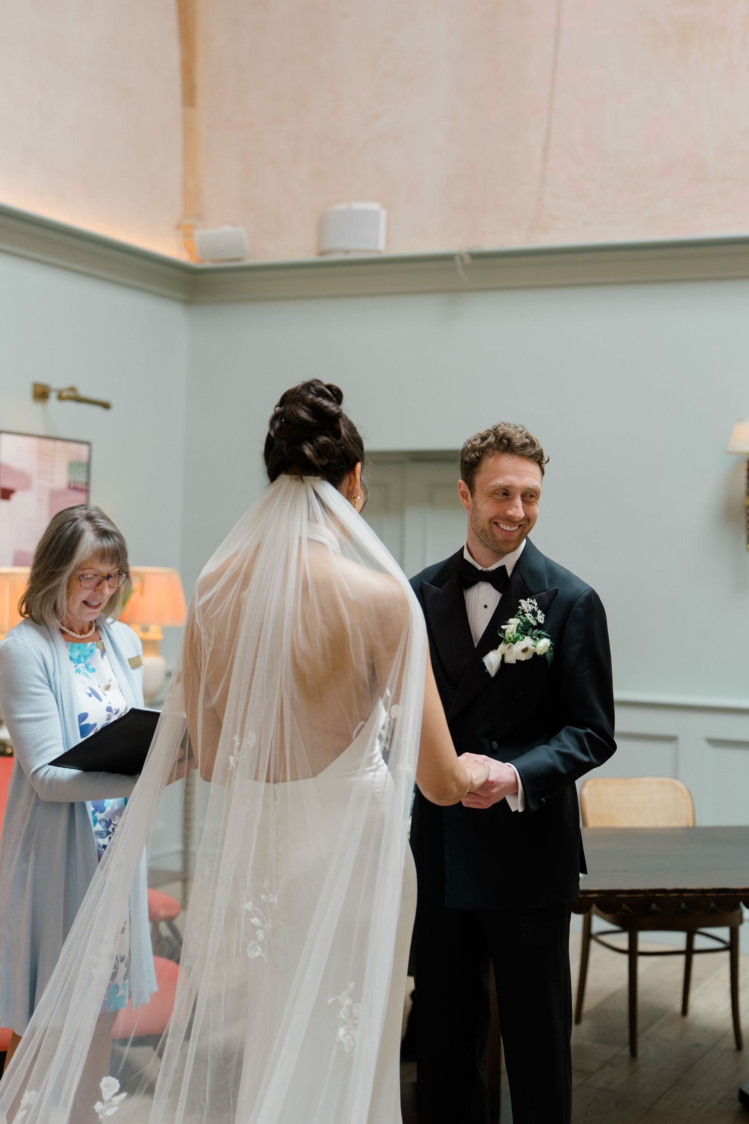 Groom smiling warmly at bride while holding hands during indoor wedding ceremony with officiant.