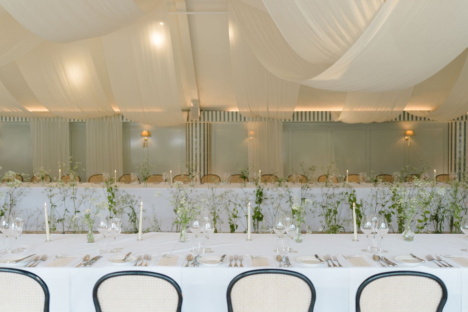 Elegant wedding reception marquee with long white tables, floral centerpieces, and draped ceiling decor.