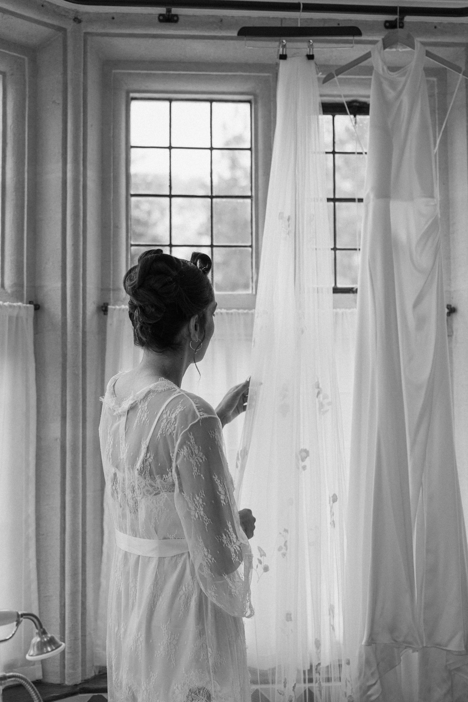 Bride in lace robe examining wedding veil hanging by window in black and white photo.