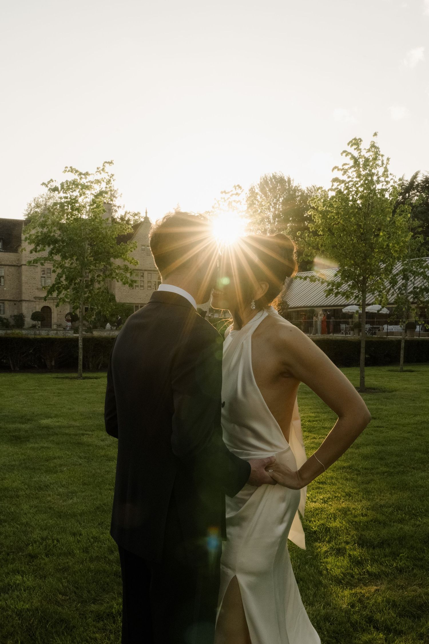 Bride and groom silhouetted against golden sunset with dramatic lens flare in garden setting.