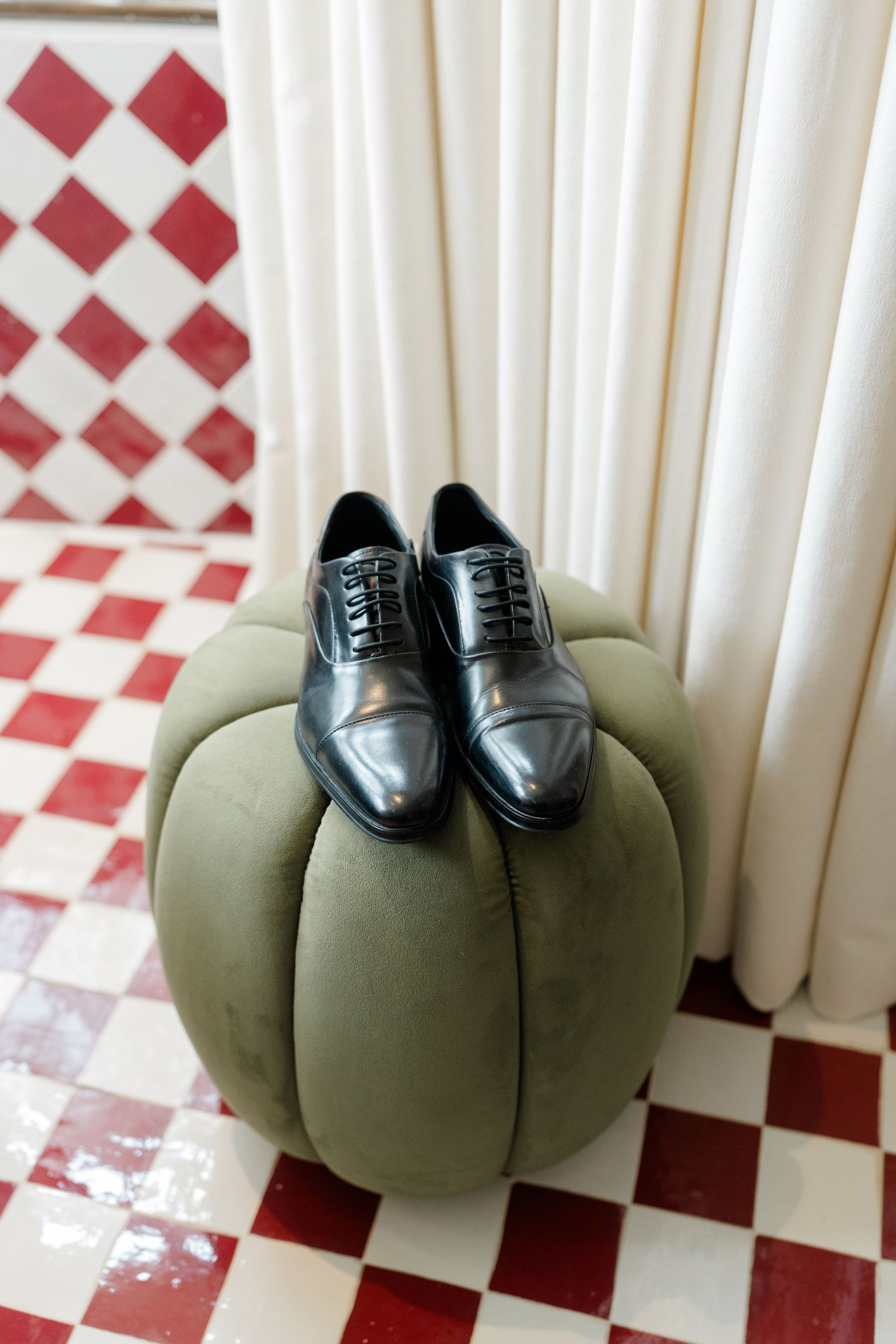 Polished black leather oxford dress shoes with laces rest on a round green velvet ottoman over a red and white checkered floor.