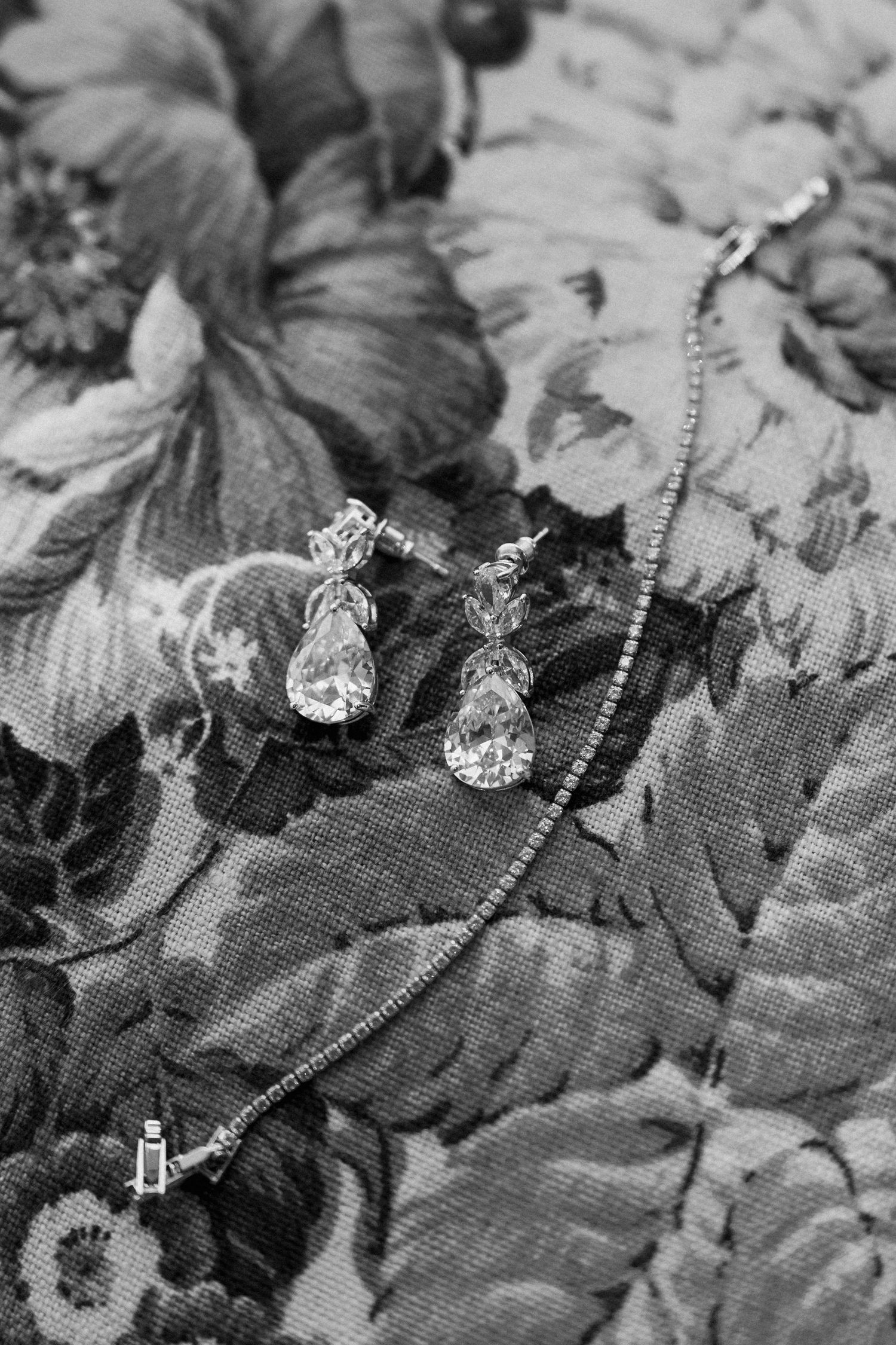 Black and white photo of teardrop crystal earrings and delicate diamond bracelet on floral fabric.