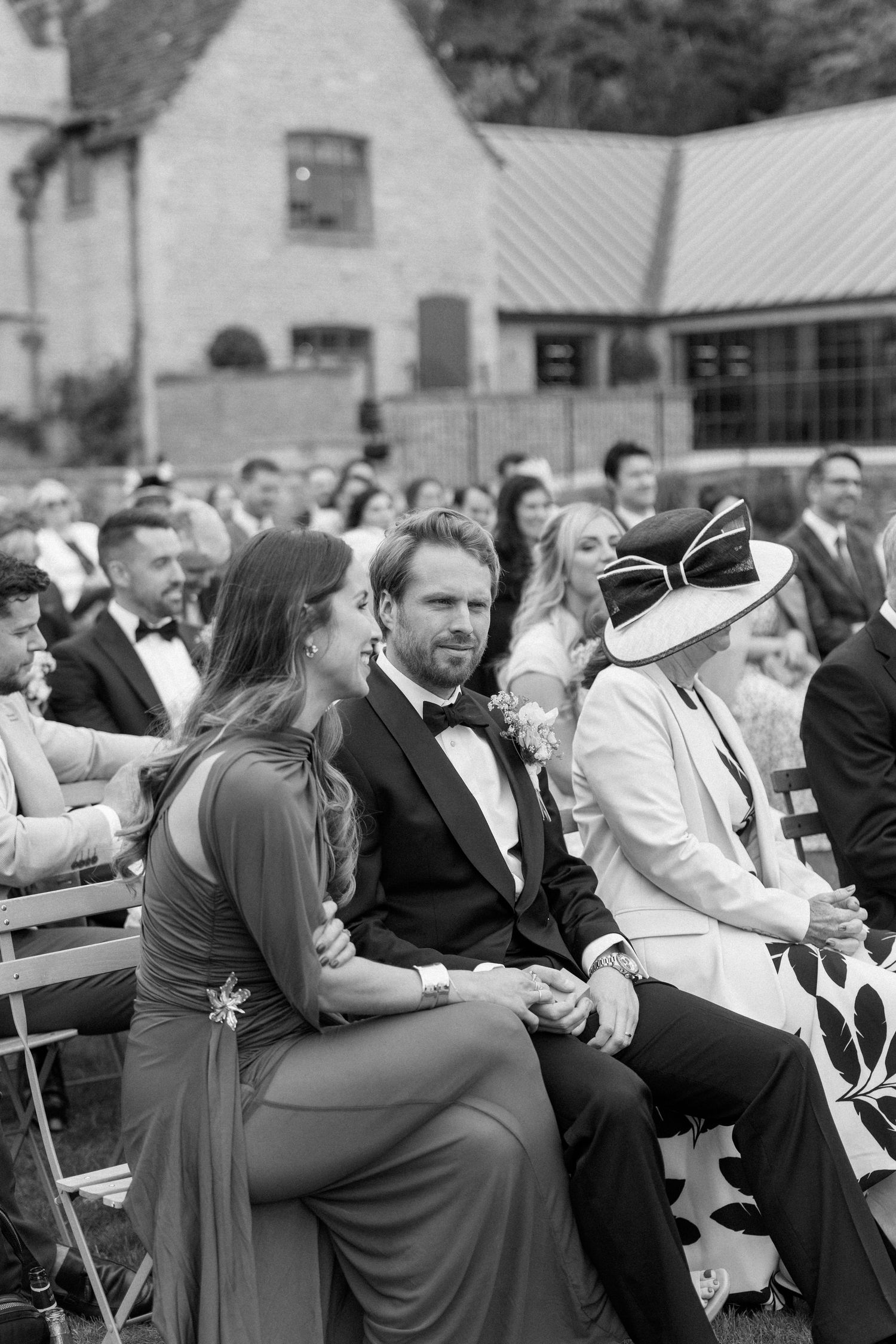 Wedding guests in formal attire seated outdoors, including woman in striking white hat at ceremony.