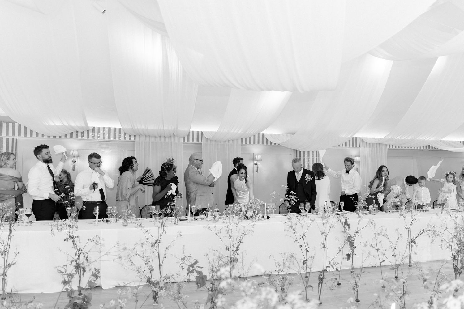 Wedding party guests celebrate at elegantly decorated head table inside draped marquee reception.