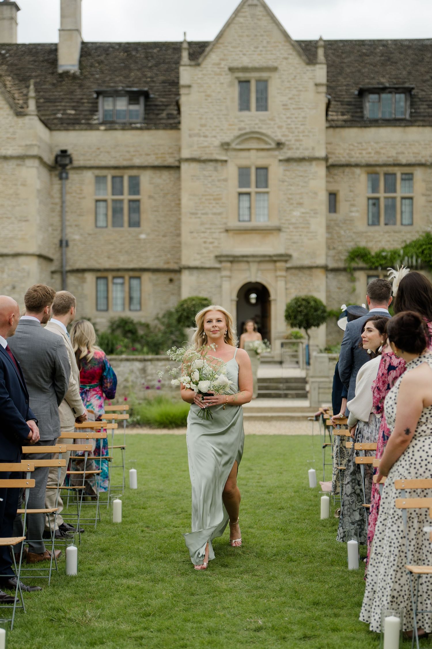 Bridesmaid in sage green dress walks down outdoor aisle of a grand English manor house wedding.