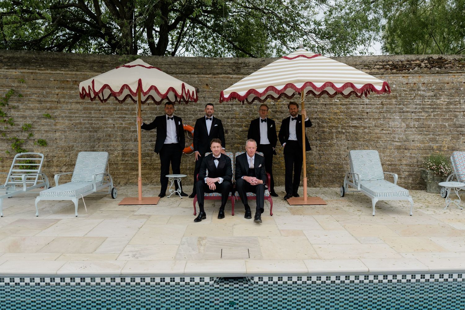 Groomsmen in black tuxedos pose poolside under striped red and white umbrellas at a luxury wedding venue.