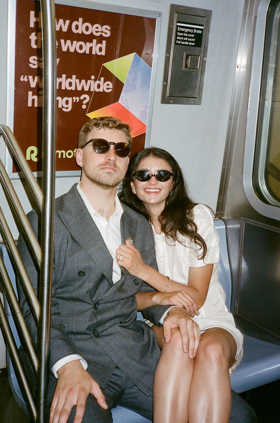 Stylish couple wearing sunglasses sitting together on a New York City subway, man in gray suit, woman in white dress.