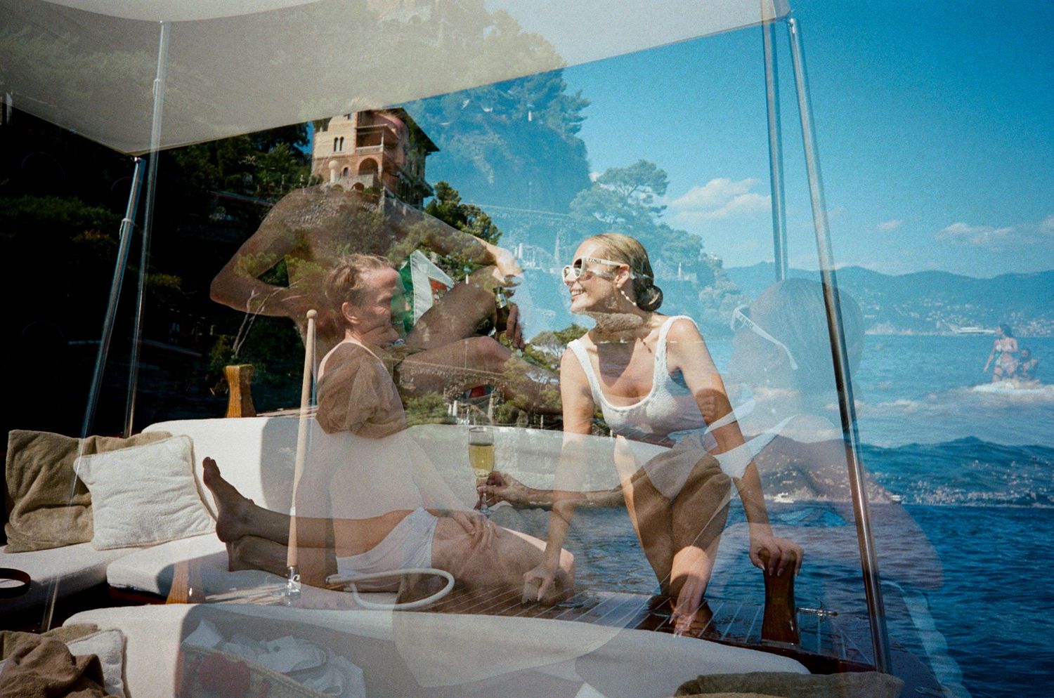 Two people relaxing on a luxury boat terrace with scenic lake and mountain views reflected in glass.