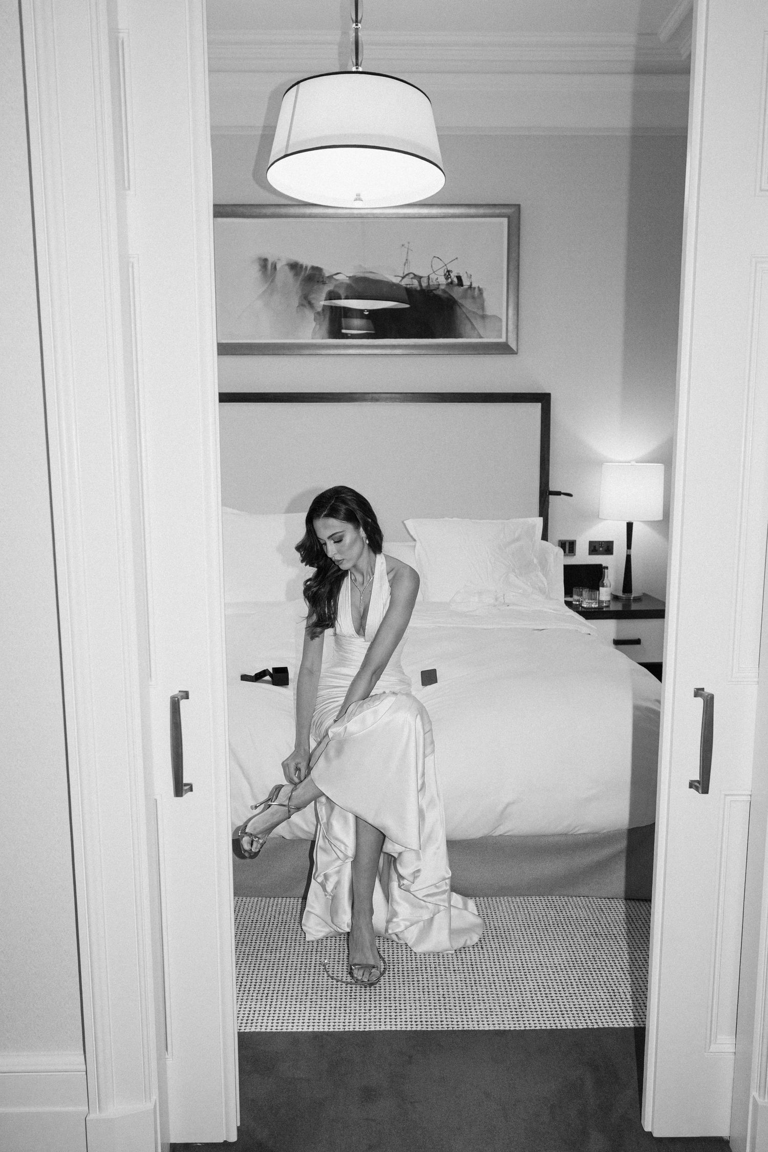 Bride in white satin gown fastening sandals in elegant hotel room, black and white wedding photo.