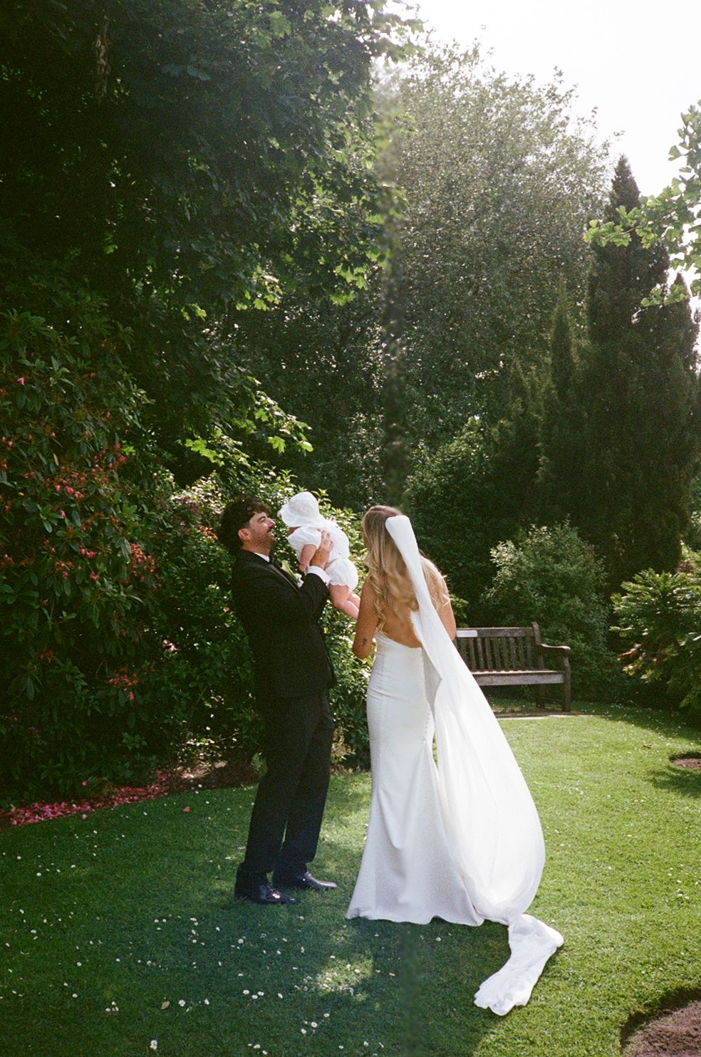 Bride and groom in garden, groom lifting baby in air, lush greenery backdrop, wedding attire.