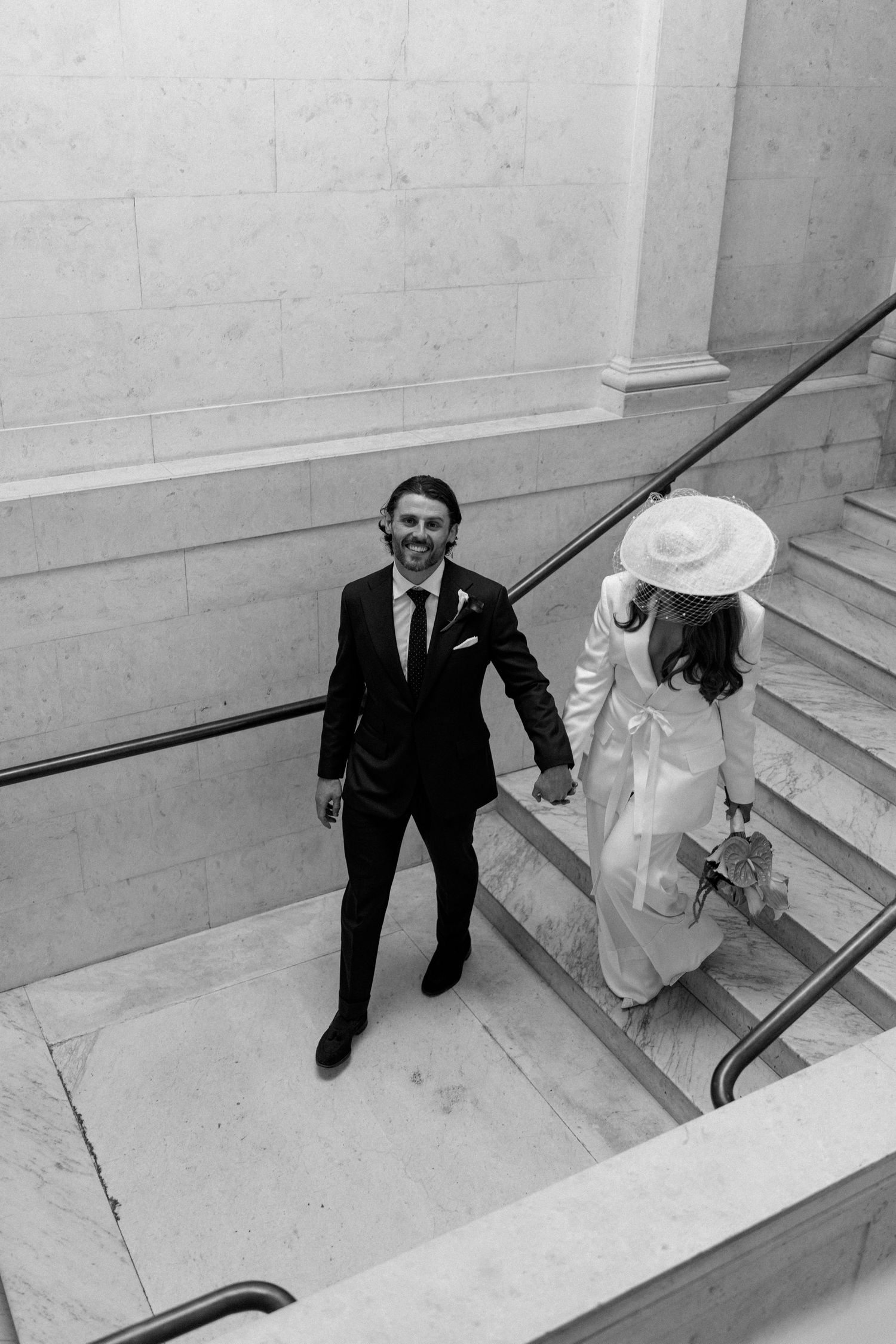 Black and white photo of smiling groom holding bride's hand on marble staircase.