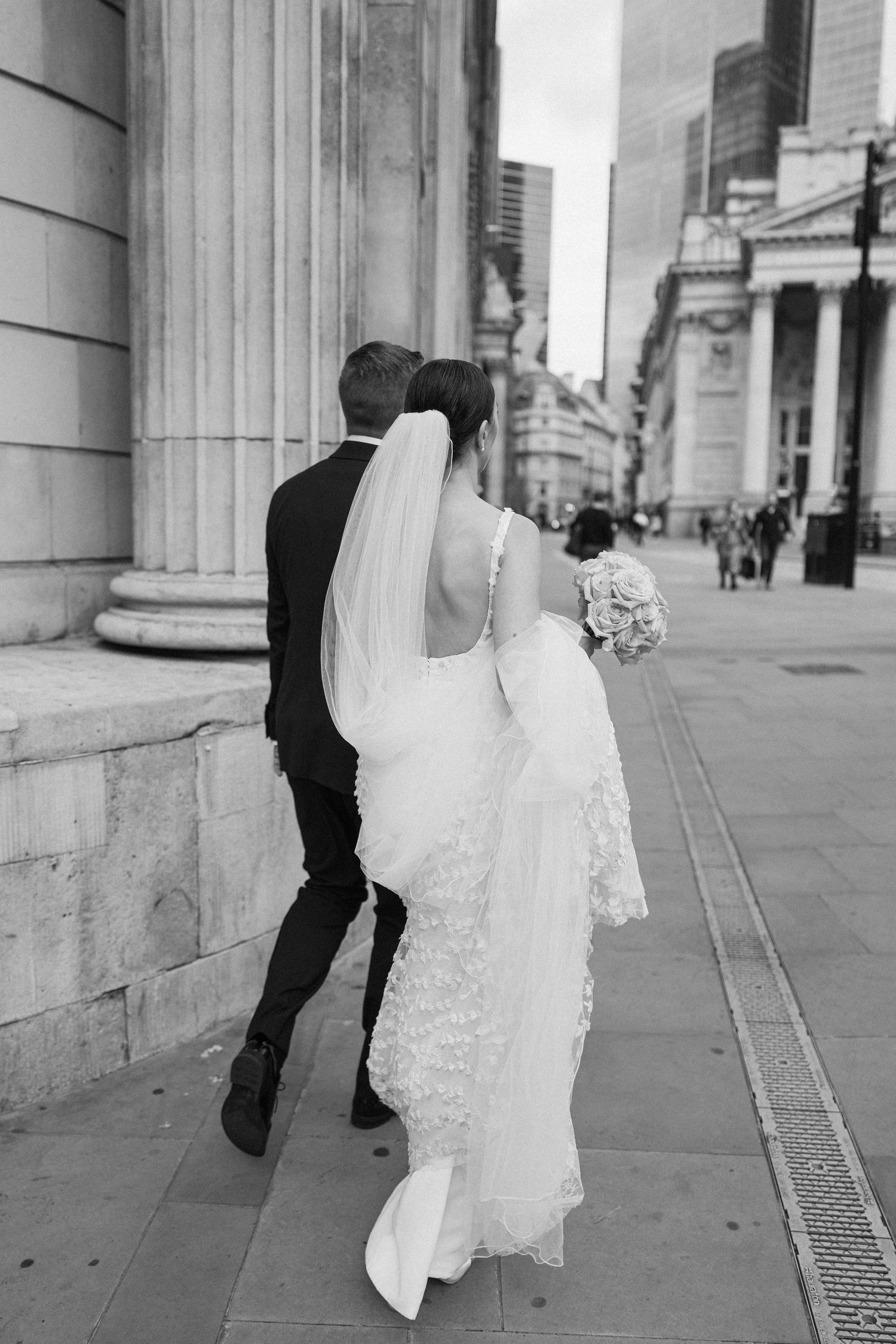 Black and white photo of a couple in wedding attire walking past classical columns.