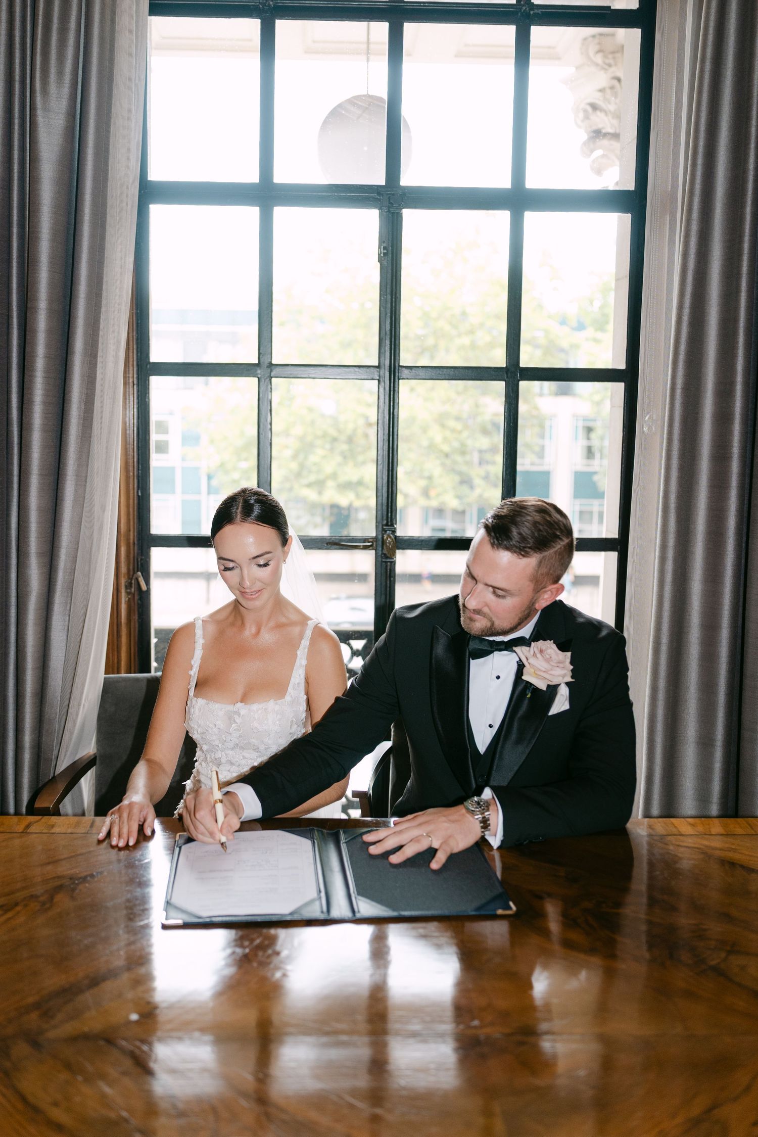 Wedding party members gather around a wooden table in a bright room with large windows.