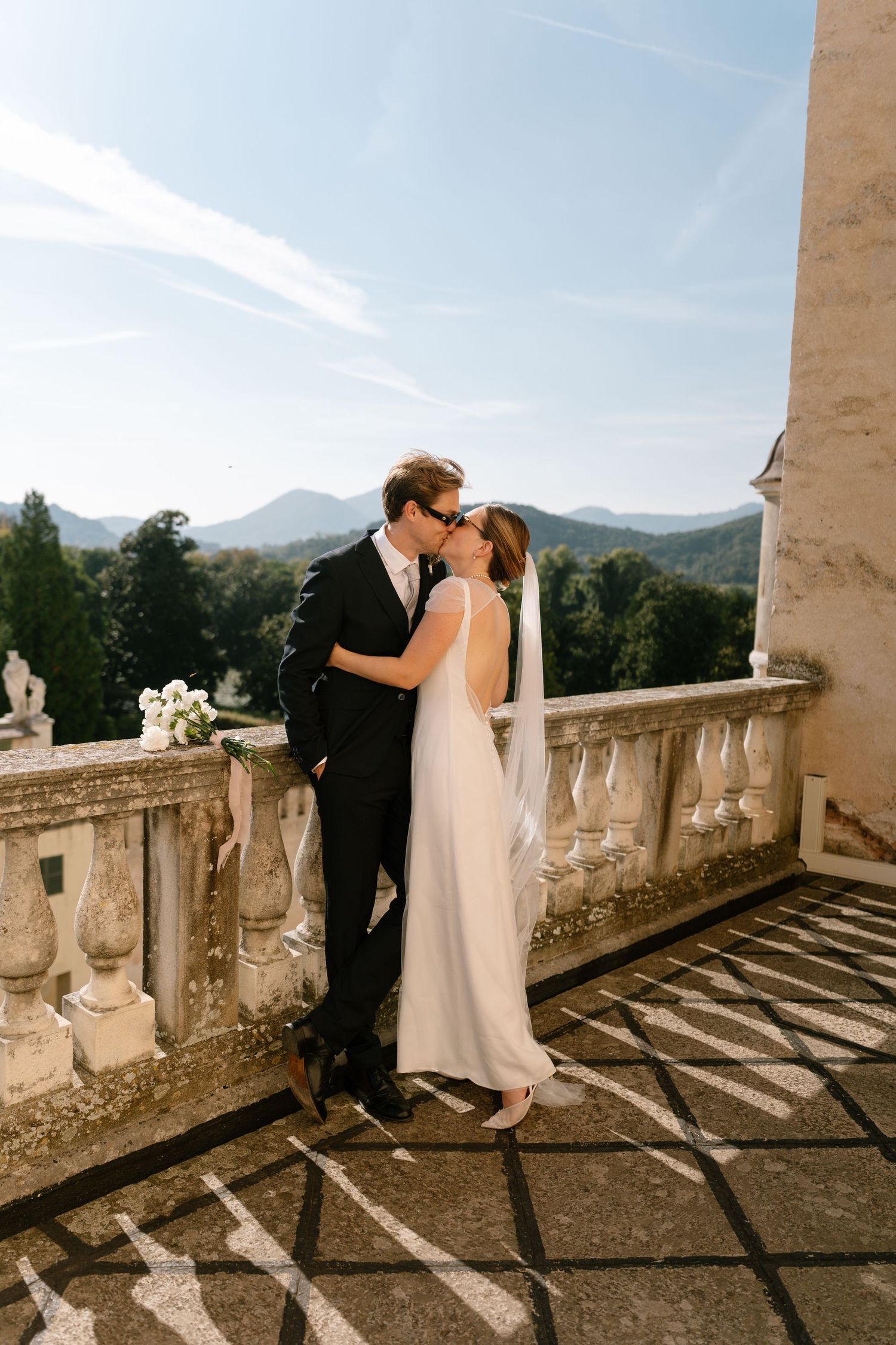 A romantic wedding couple shares a kiss on an elegant stone balcony overlooking mountain views.
