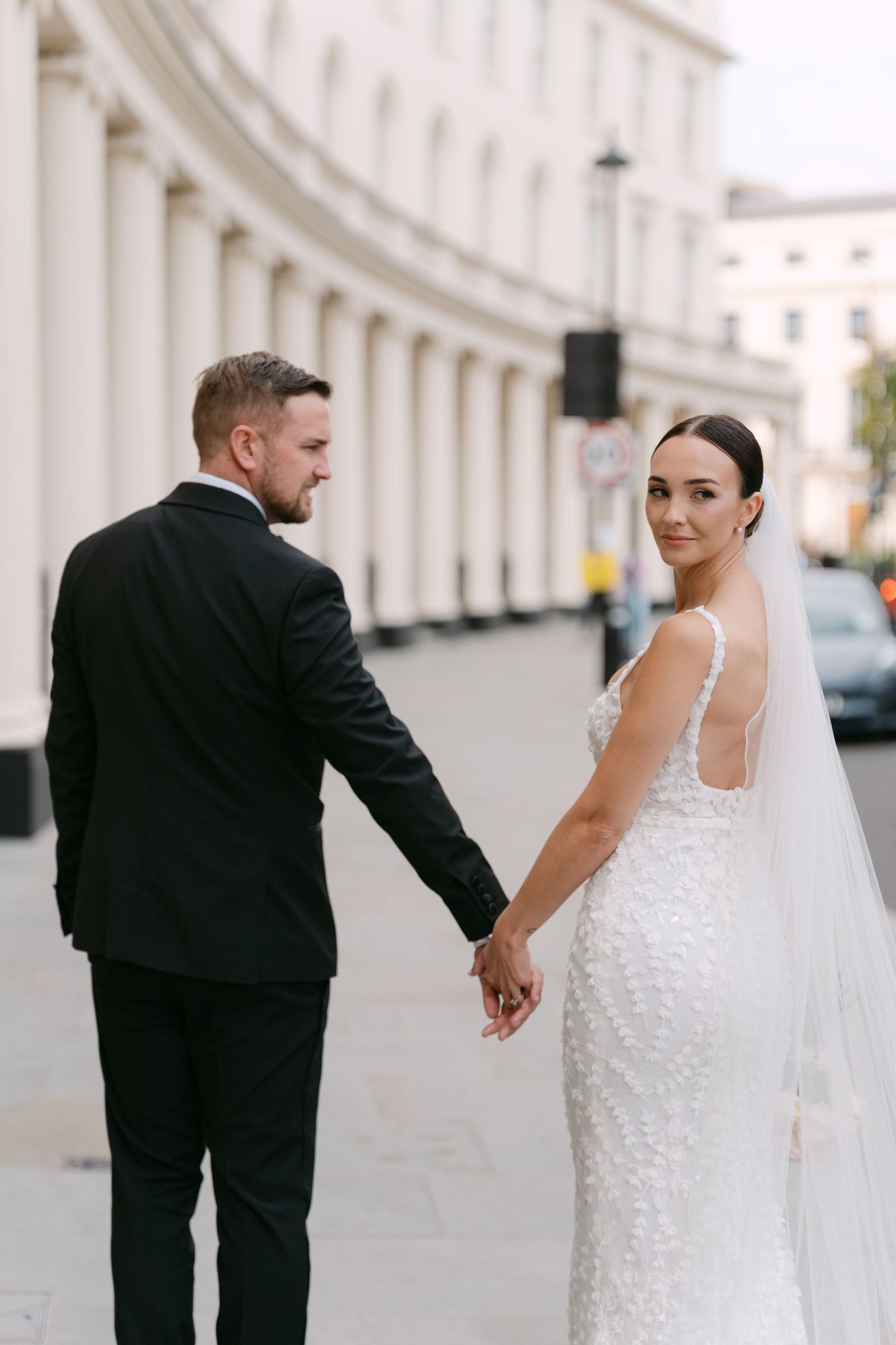 A couple in wedding attire holds hands while walking along a curved colonnade.