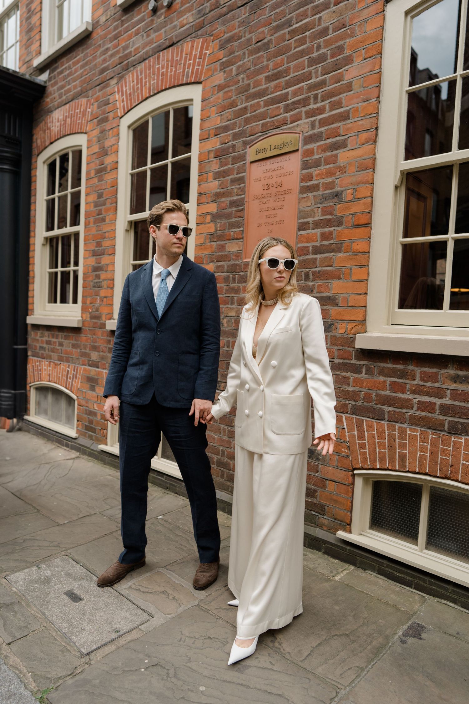 Stylish couple in sunglasses pose against brick building, wearing navy suit and white pantsuit.