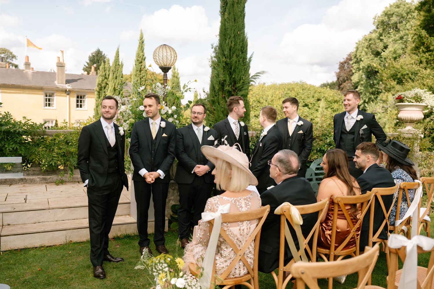 Wedding party gathers outdoors at an elegant garden ceremony with wooden chairs and cypress trees.