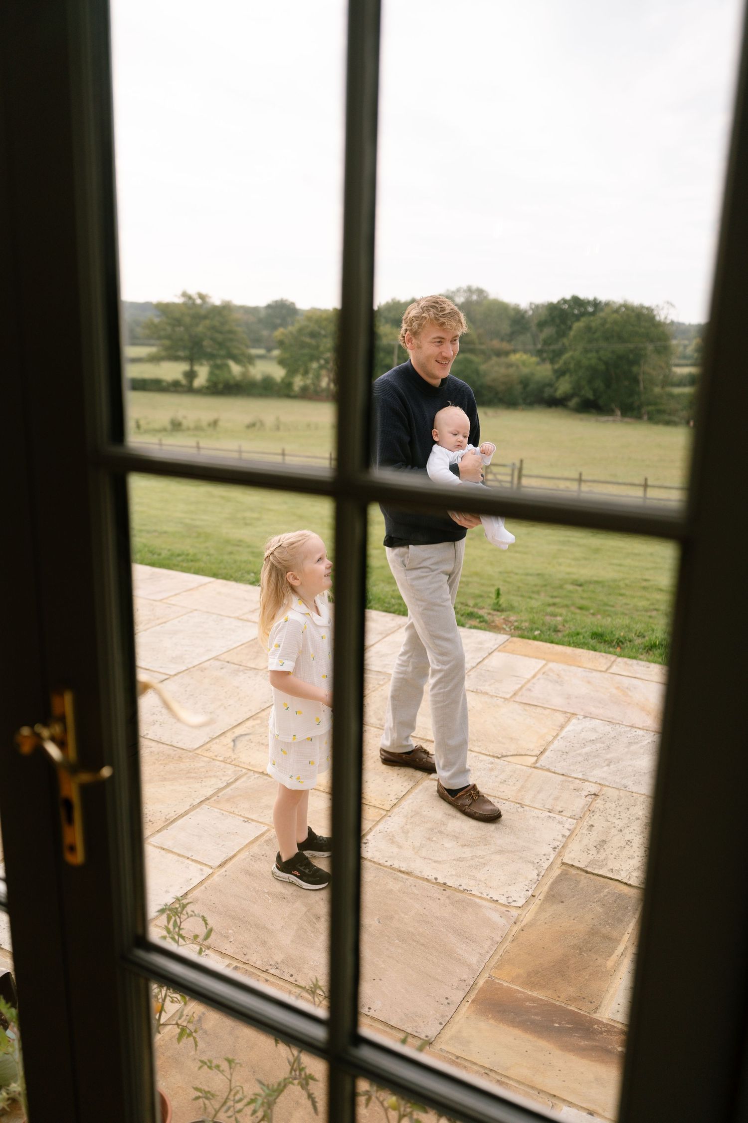 View through window panes of people standing on a stone patio overlooking countryside fields.