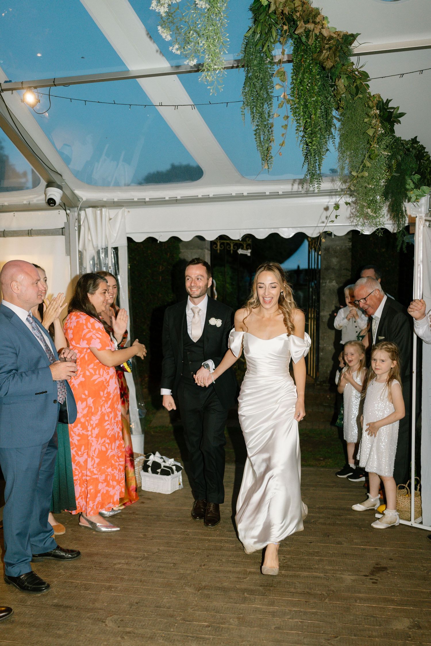 A couple makes their grand entrance at a wedding reception under a tent with hanging greenery and string lights.