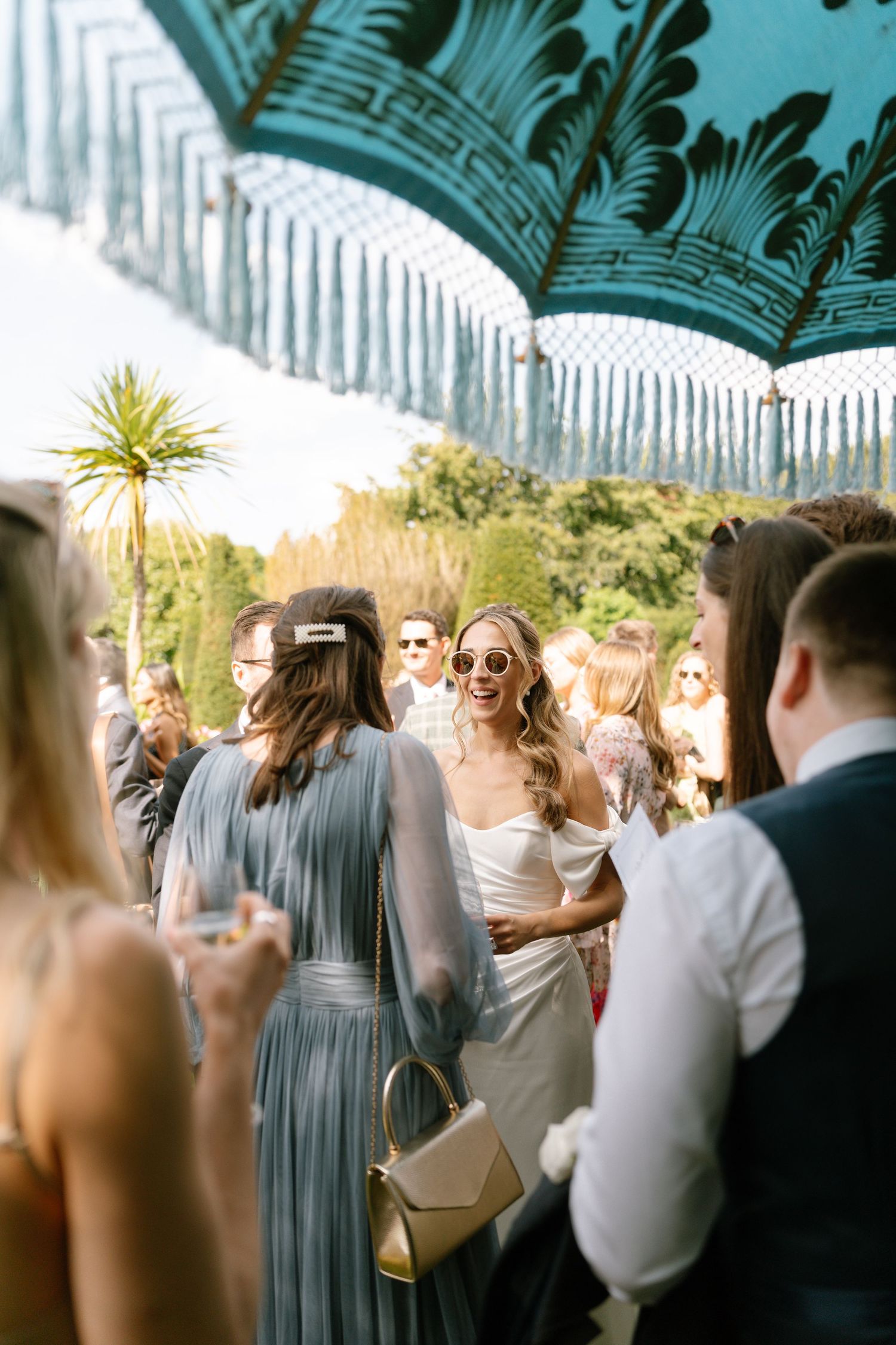 Guests mingle under a decorative turquoise umbrella at an elegant outdoor garden party.