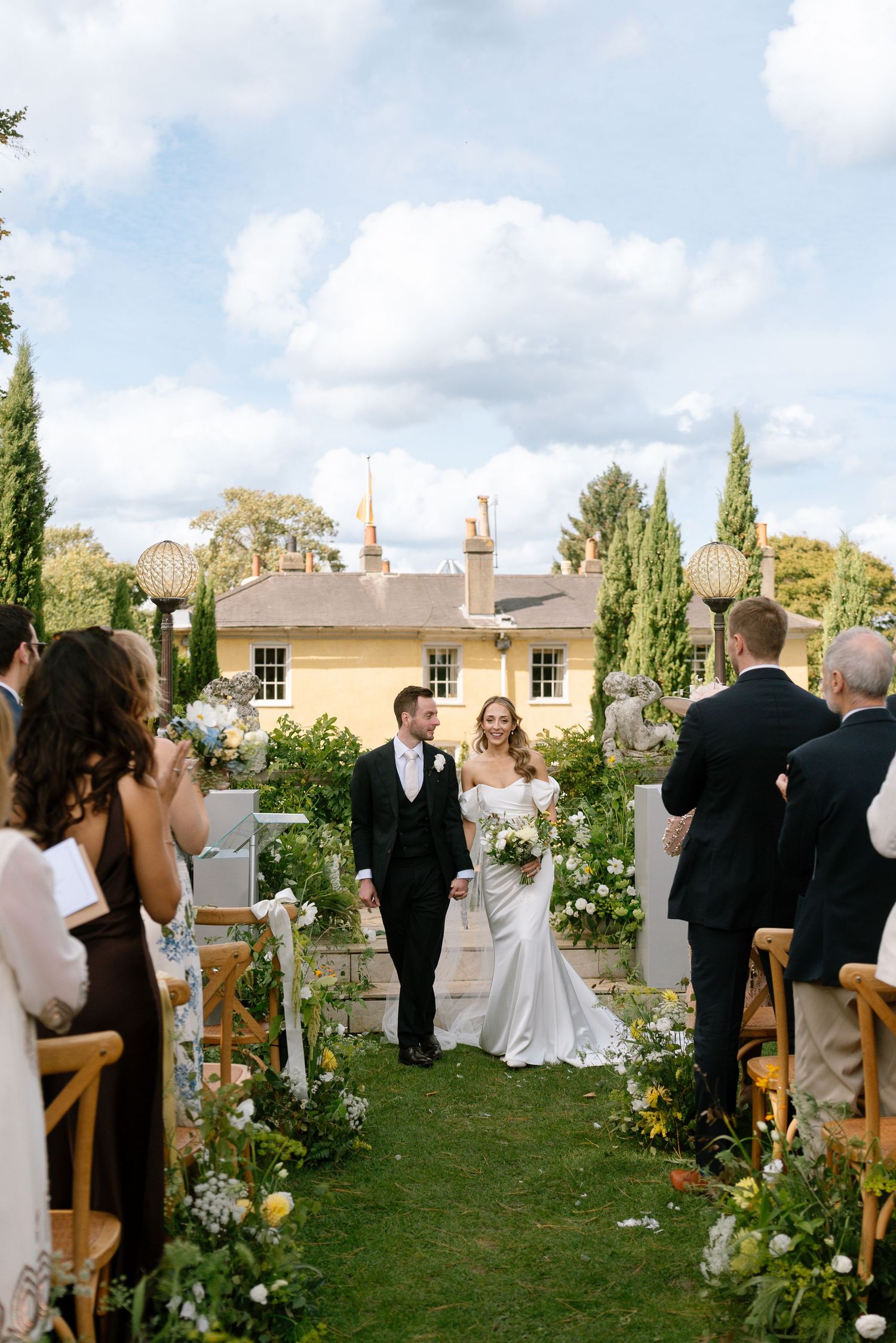 Wedding guests stand during ceremony in garden setting with cypress trees and historic building.