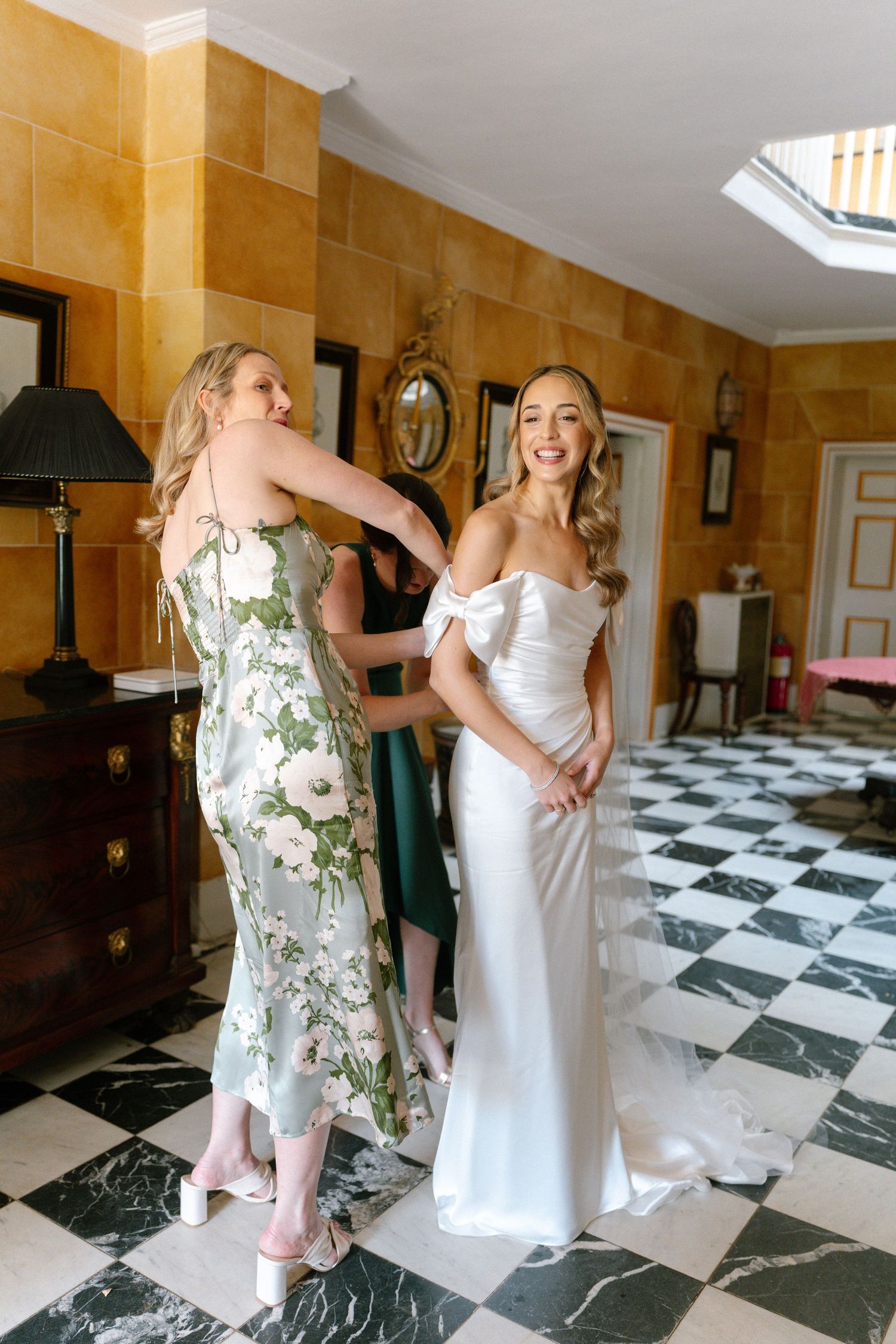 Two women in elegant dresses share a joyful moment on black and white checkered floor.