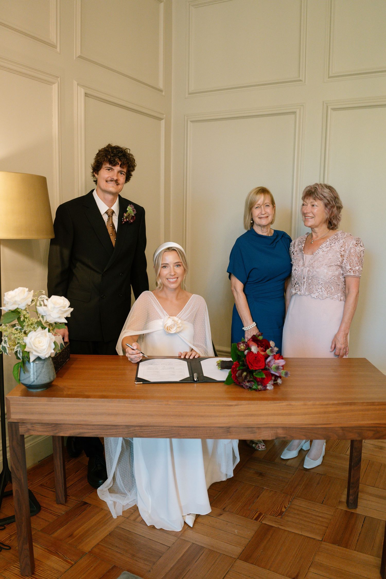 Wedding party gathered around a wooden table for document signing with white floral arrangements.