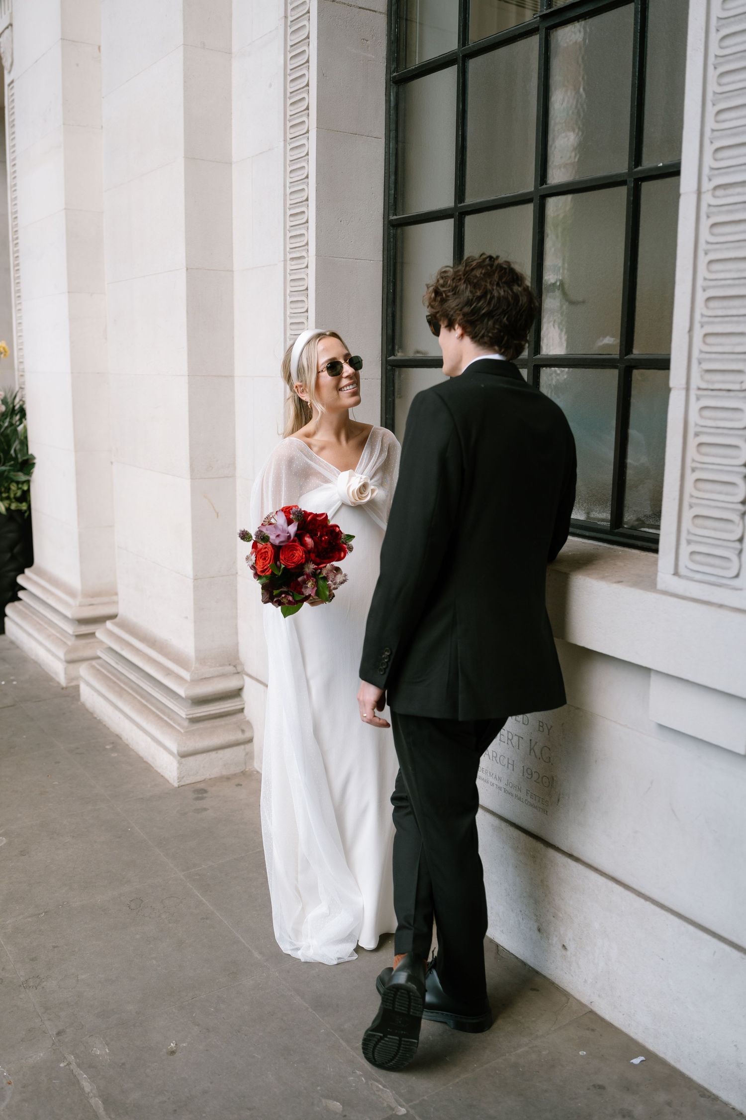 A stylish bride in sunglasses holds red flowers while talking with groom by ornate columns.