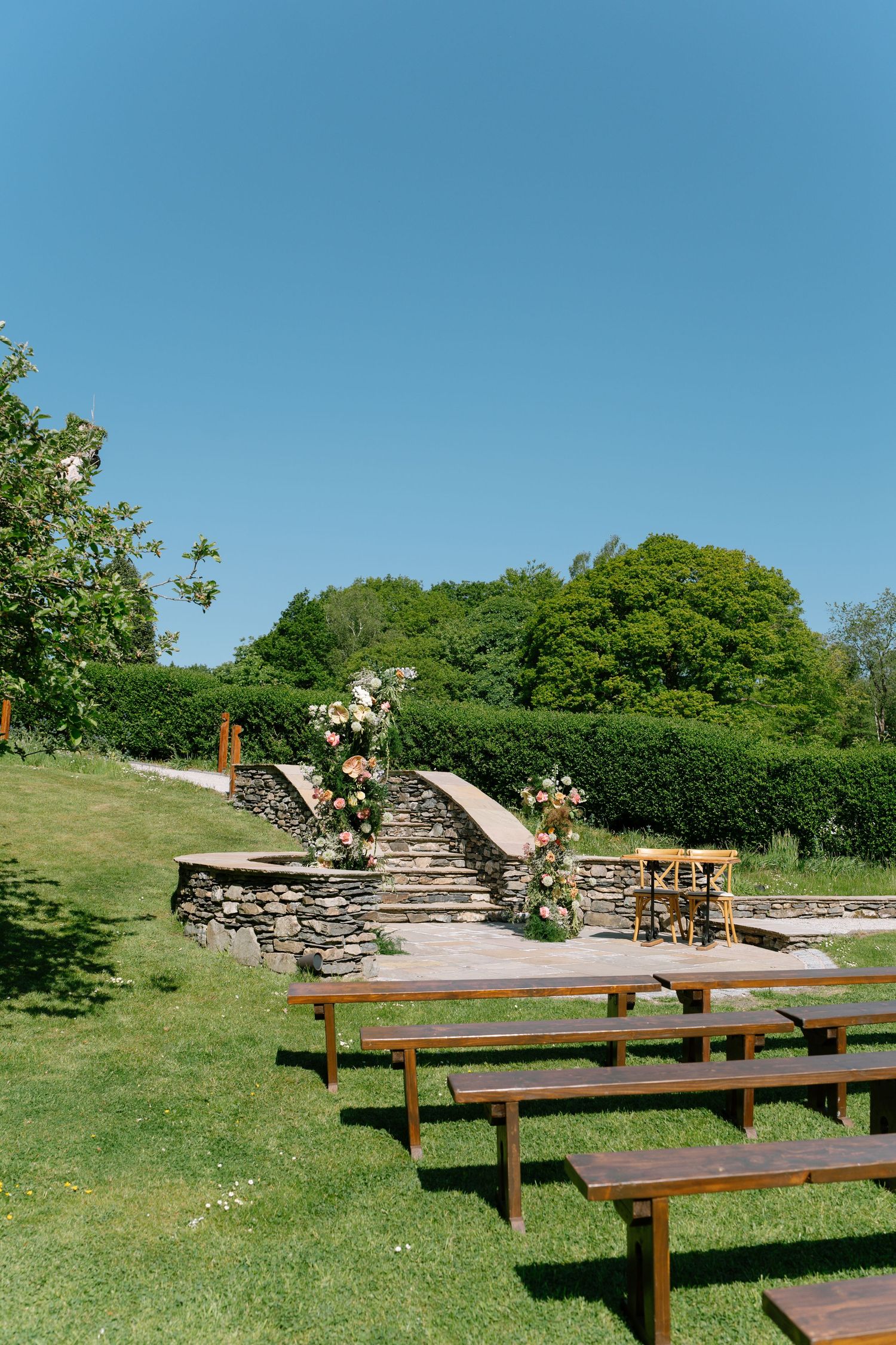 Rustic outdoor wedding ceremony setup with wooden benches and stone walls.