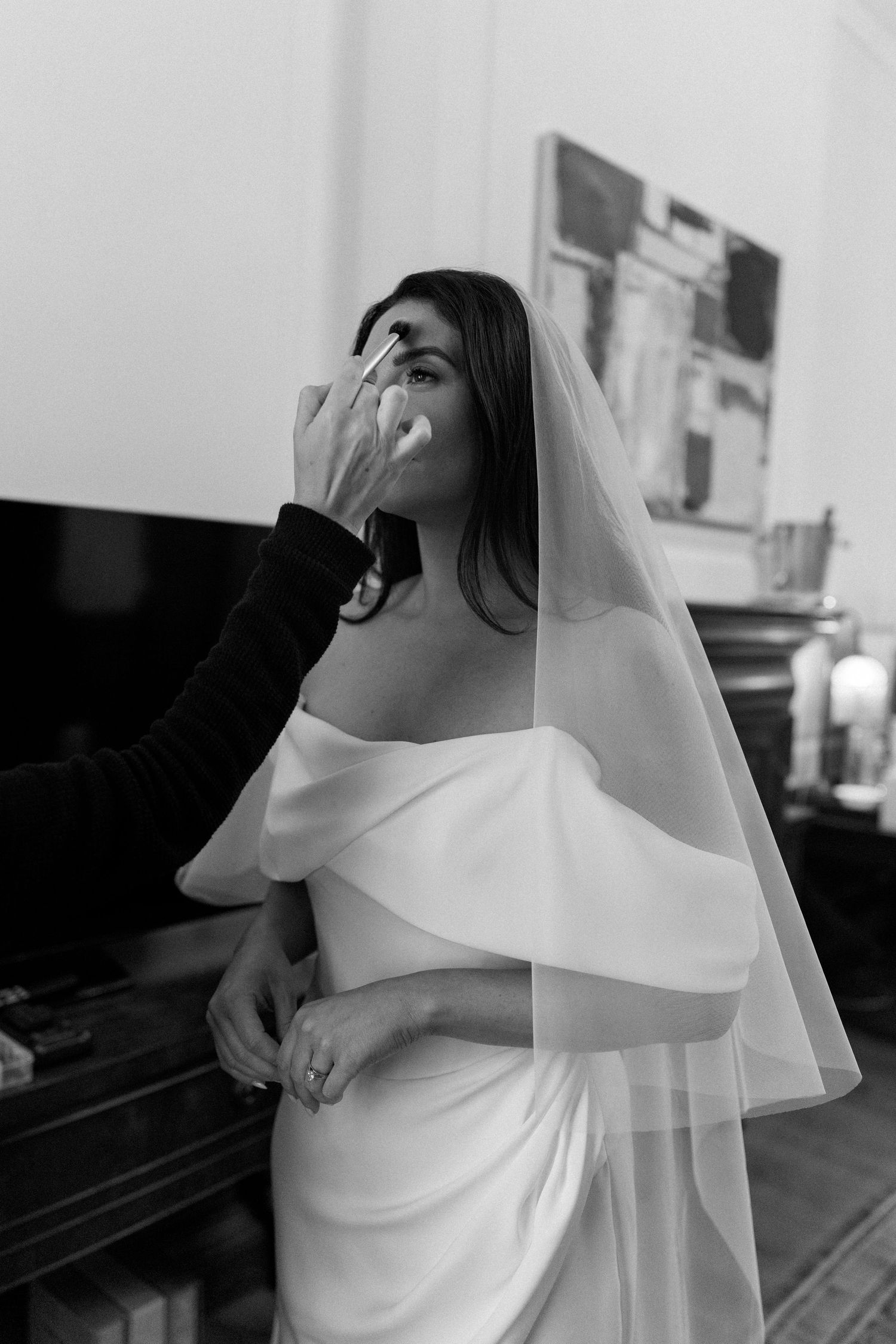 Black and white photo of a bride in an off-the-shoulder satin wedding gown and veil while a makeup artist applies her makeup.
