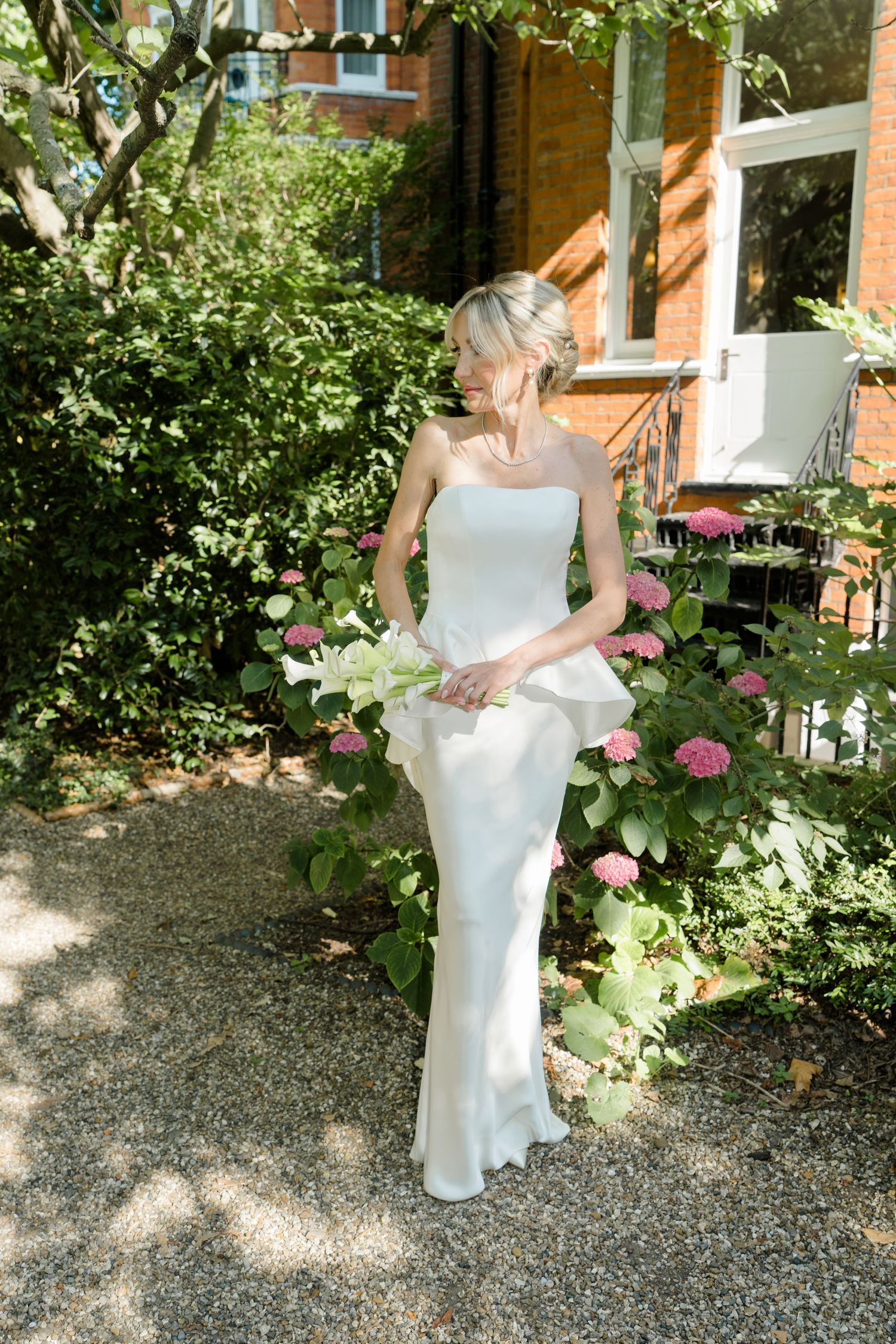 Bride in white strapless gown holding flowers, standing in a lush garden with pink hydrangeas and brick building.