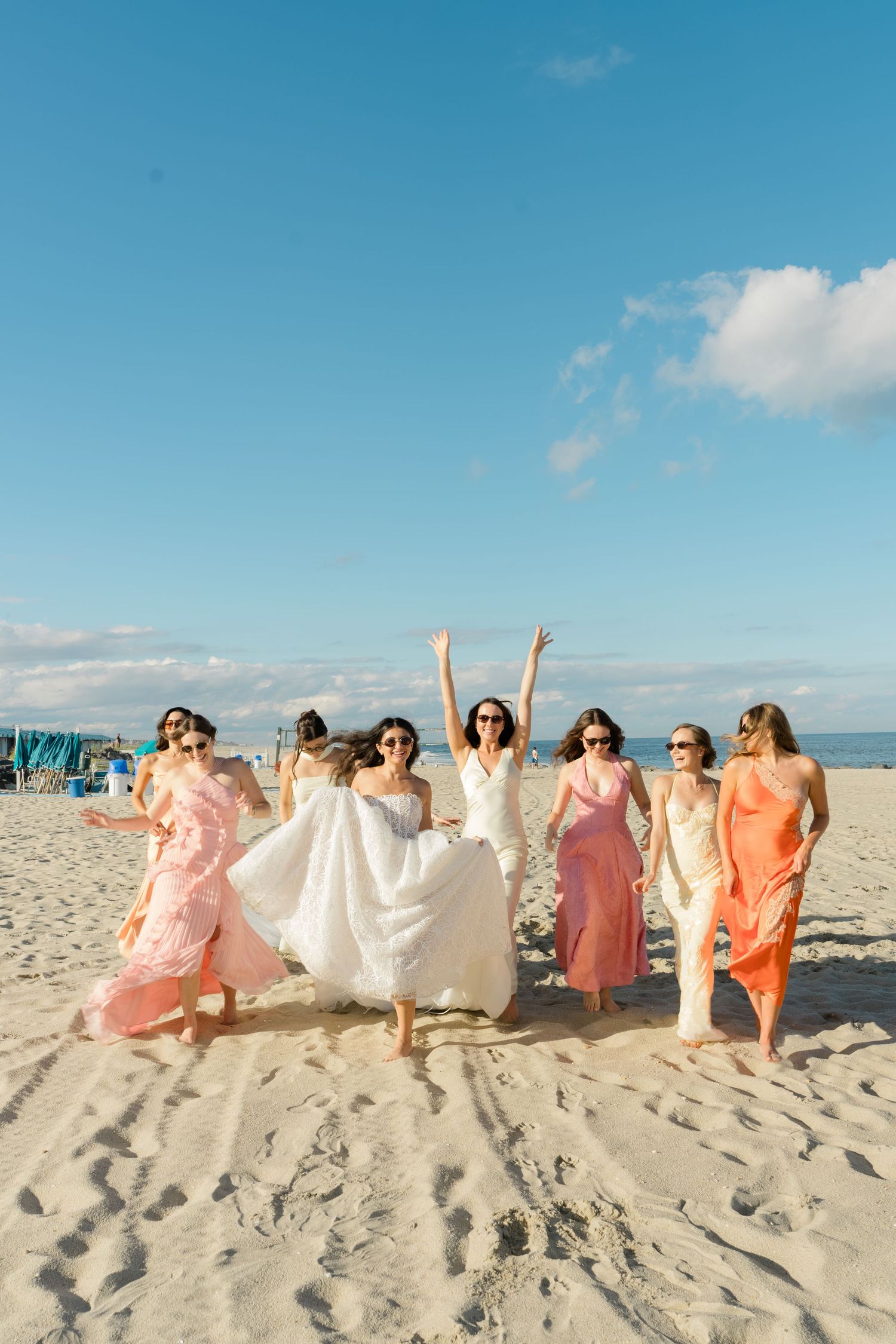 Bride in white dress jumping joyfully with bridesmaids in colorful gowns on a sunny sandy beach.
