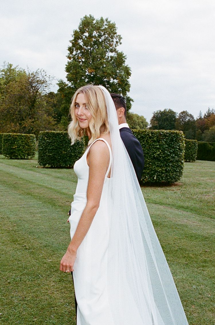 Bride in white gown and veil looks back over shoulder at groom in navy suit on lush green lawn.