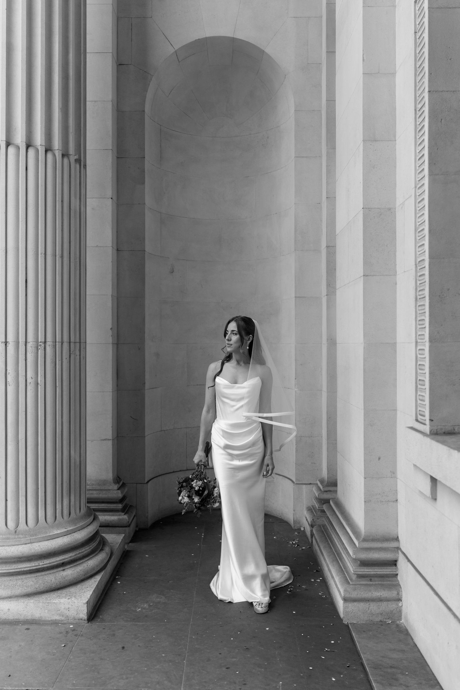 Elegant bride in white satin gown standing between stone columns.