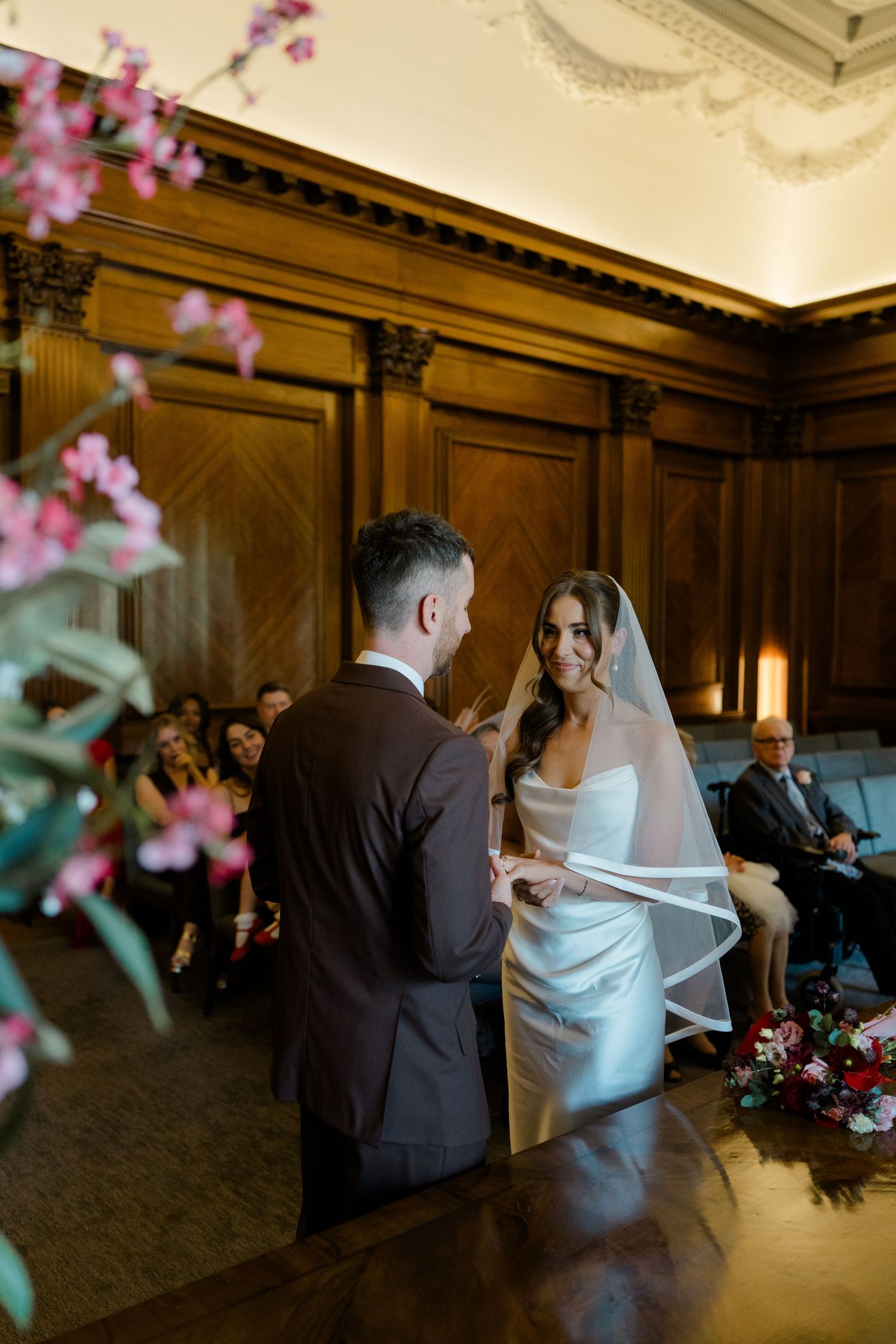 Bride smiles at groom during wedding ring exchange ceremony in elegant wood-paneled room with floral decor.