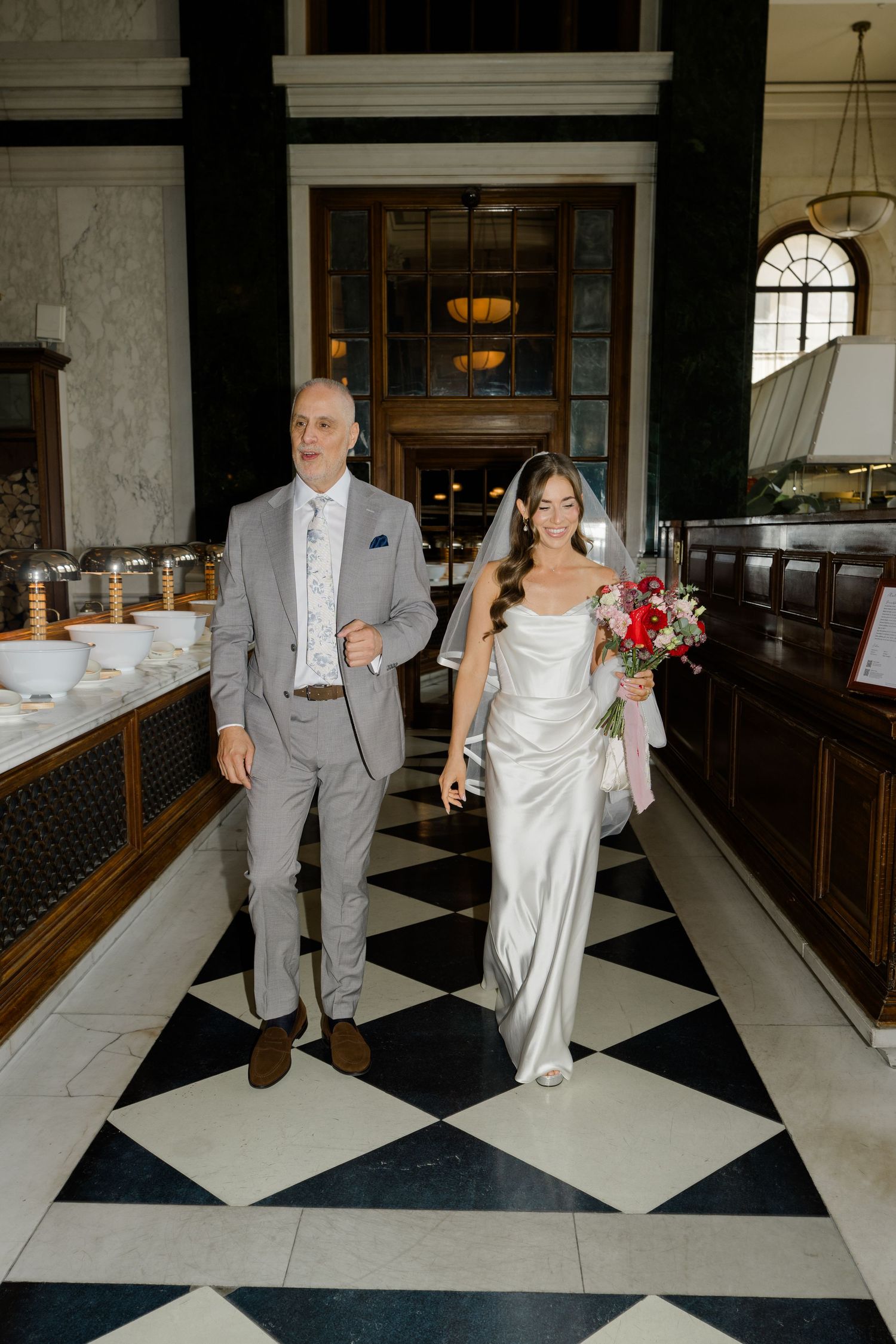 A bride in a satin slip gown holding a red bouquet walks the aisle with her father in a gray suit.